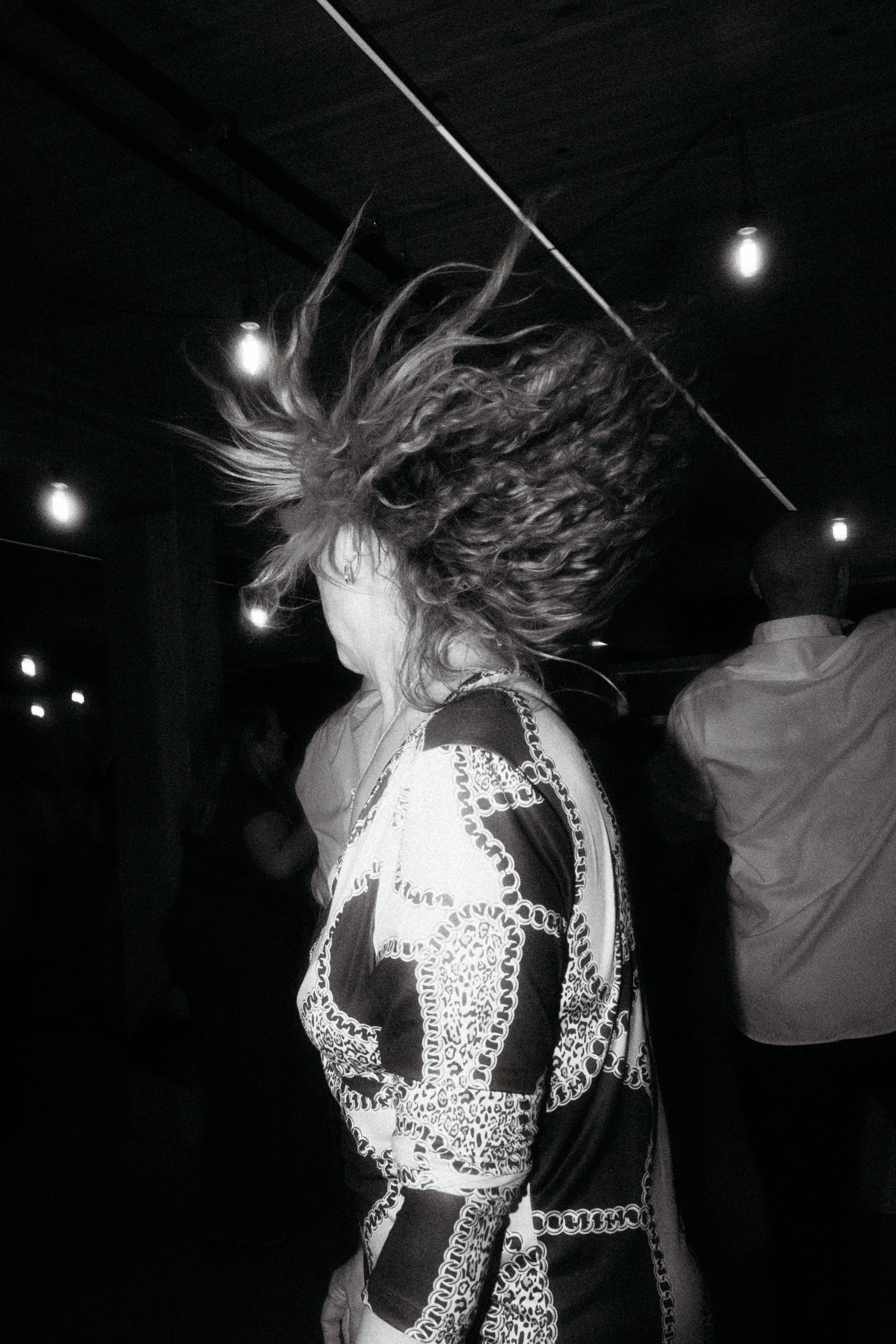 A woman with curly hair flipping her hair at a dance or party, in black and white. She is wearing a patterned dress or top. Other people are visible in the background in a dimly lit setting.