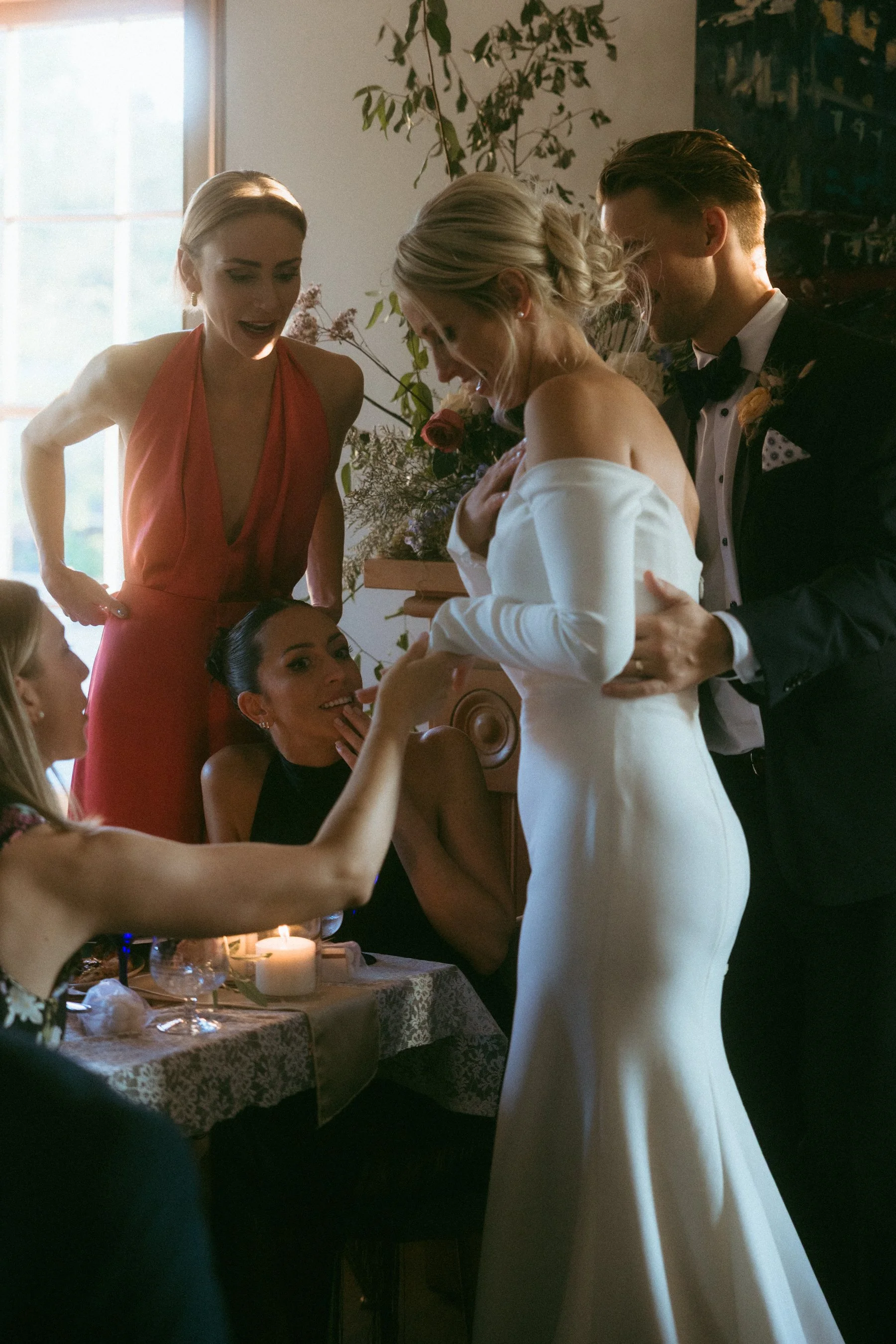 A bride in a strapless white gown is surrounded by friends, with one friend applying makeup or touching her face during a wedding celebration in a warmly lit room with floral decor.
