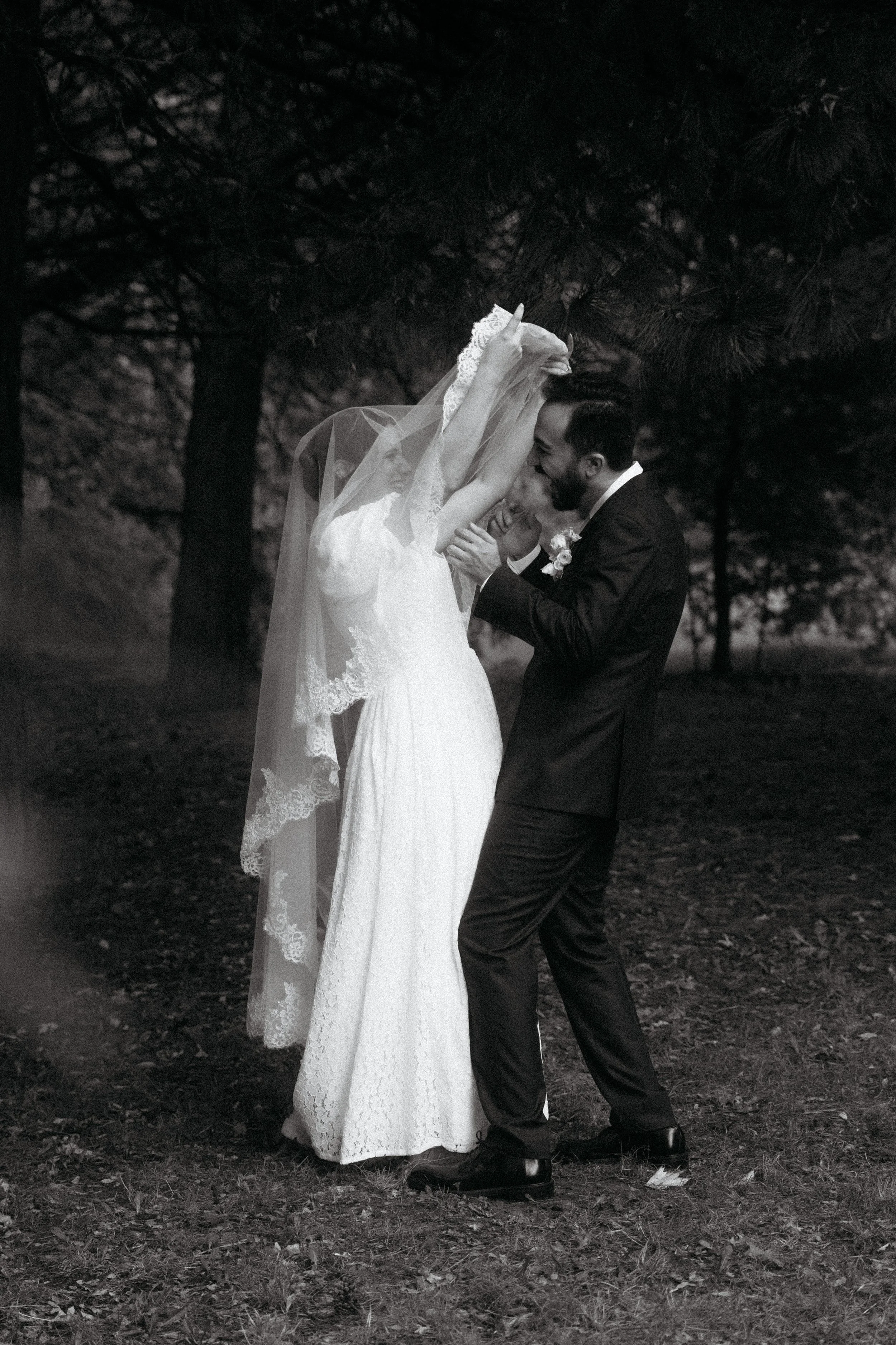 A black and white photo of a wedding scene outdoors, with a bride and groom. The bride is wearing a long lace wedding dress and veil, while the groom is in a dark suit. They are close, smiling and touching foreheads, with trees in the background.