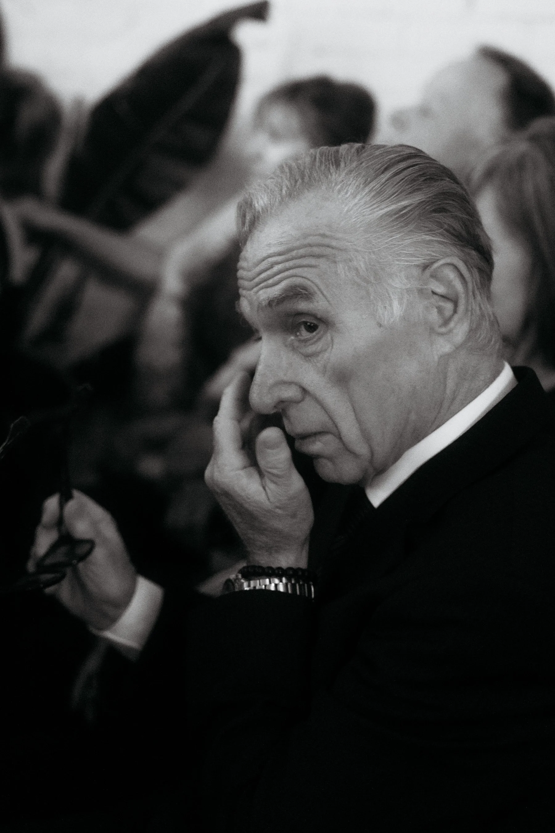 An elderly man with slicked-back hair touching his face with his left hand, holding glasses with his right hand, in a black-and-white photo at a formal event.