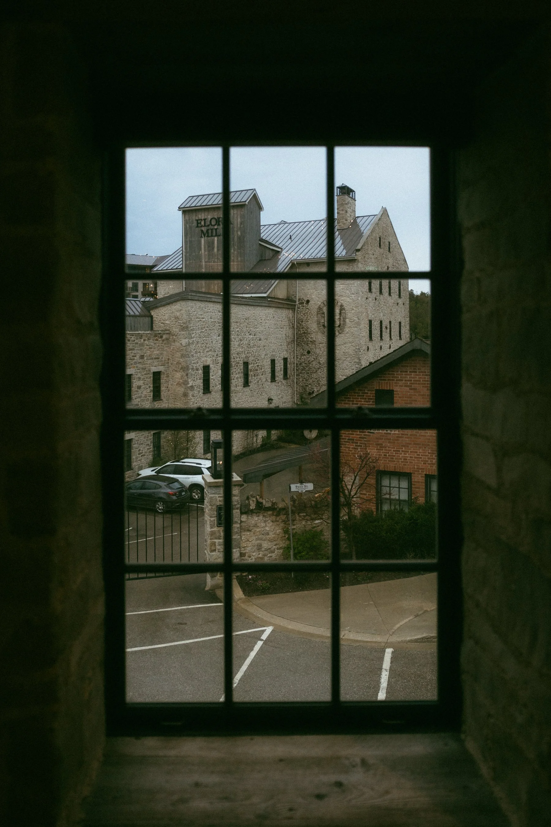 View through a small, divided window showing an historic stone building with the sign "ELOF MIL" on it, parked cars in a lot, and a brick building in the foreground.