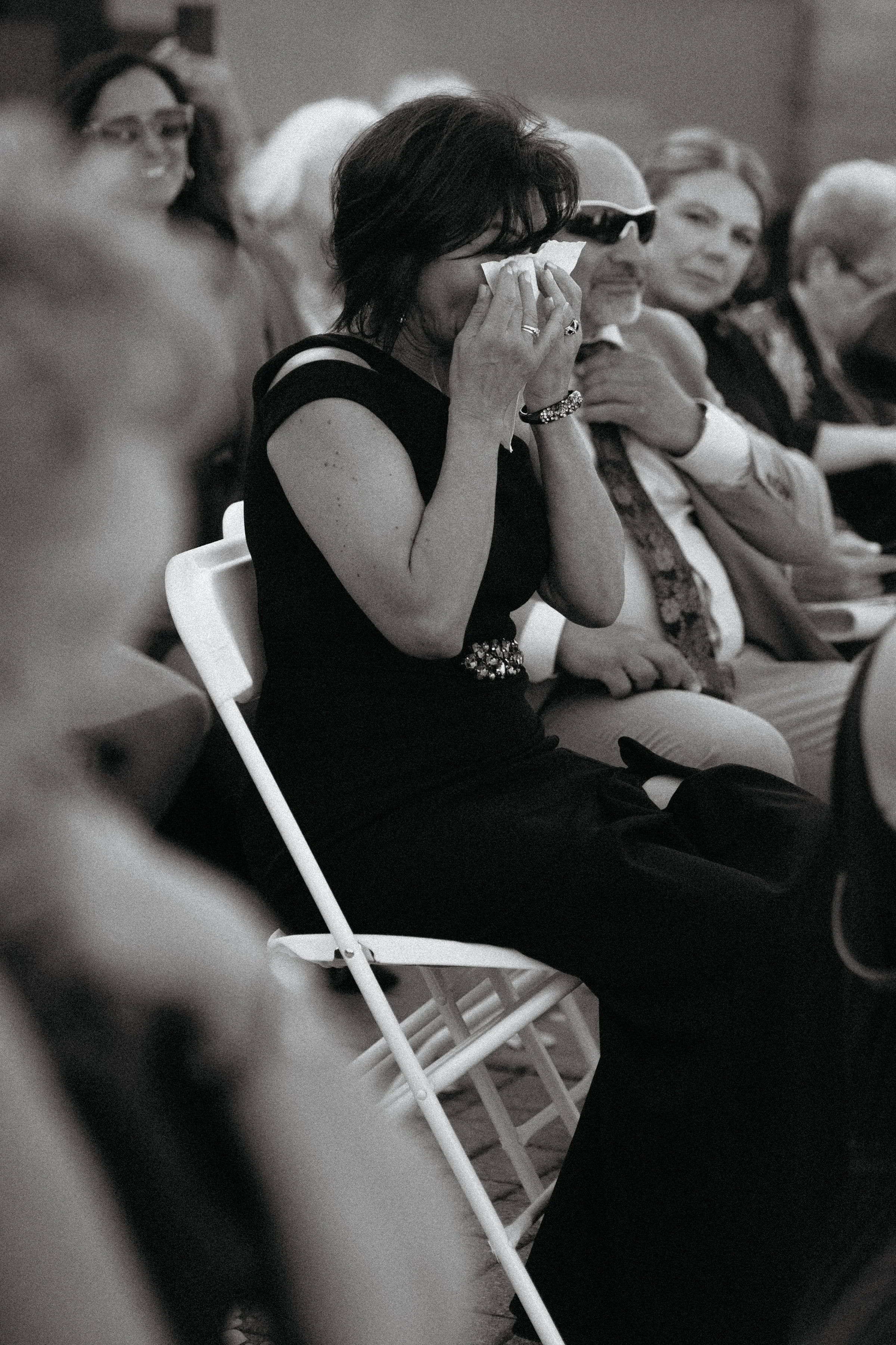 A woman in a black dress sitting on a chair, crying and wiping her face with a tissue, surrounded by other people at an indoor event.