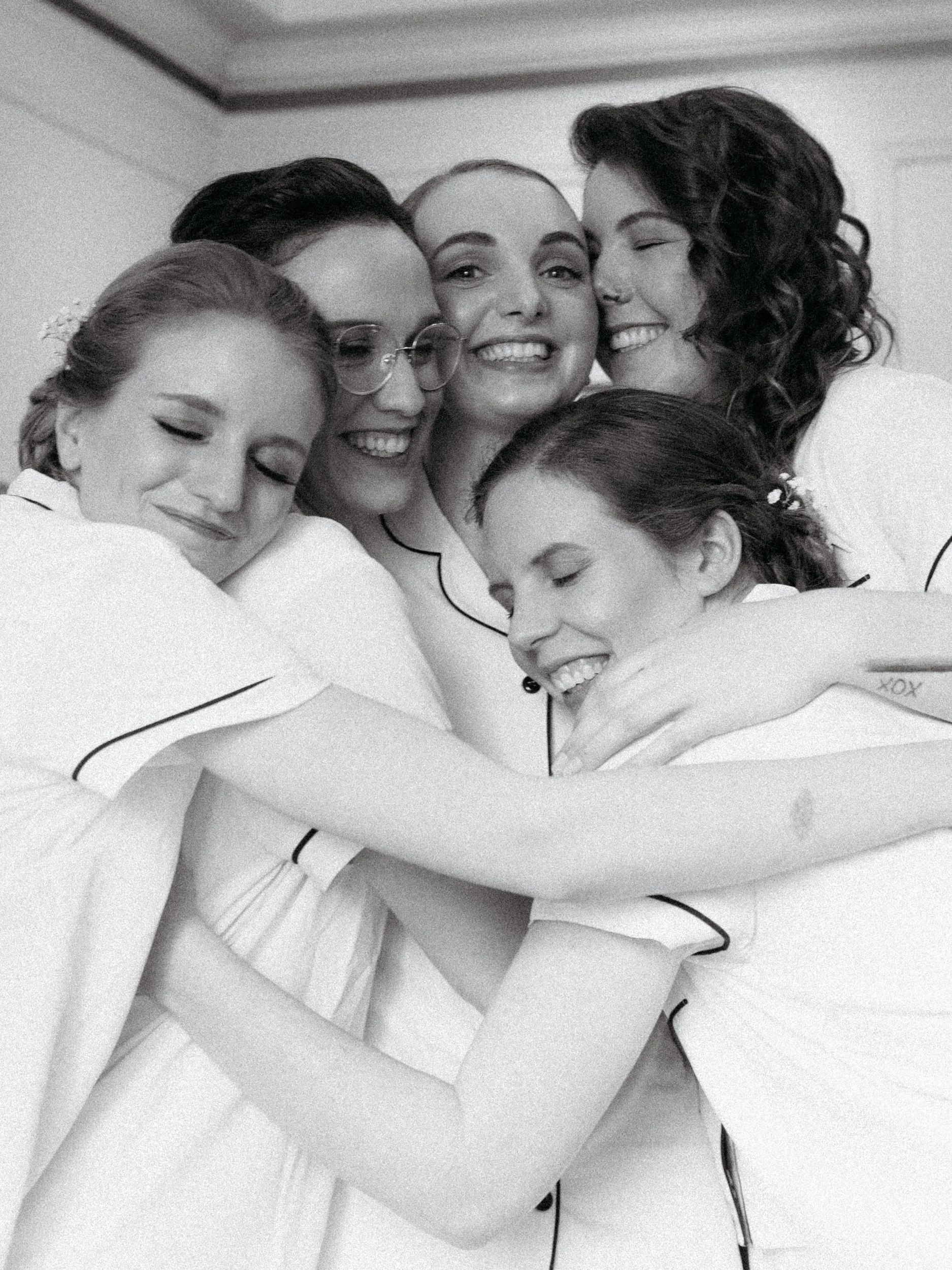 Group of five women in pajamas hugging each other and smiling, celebrating together.