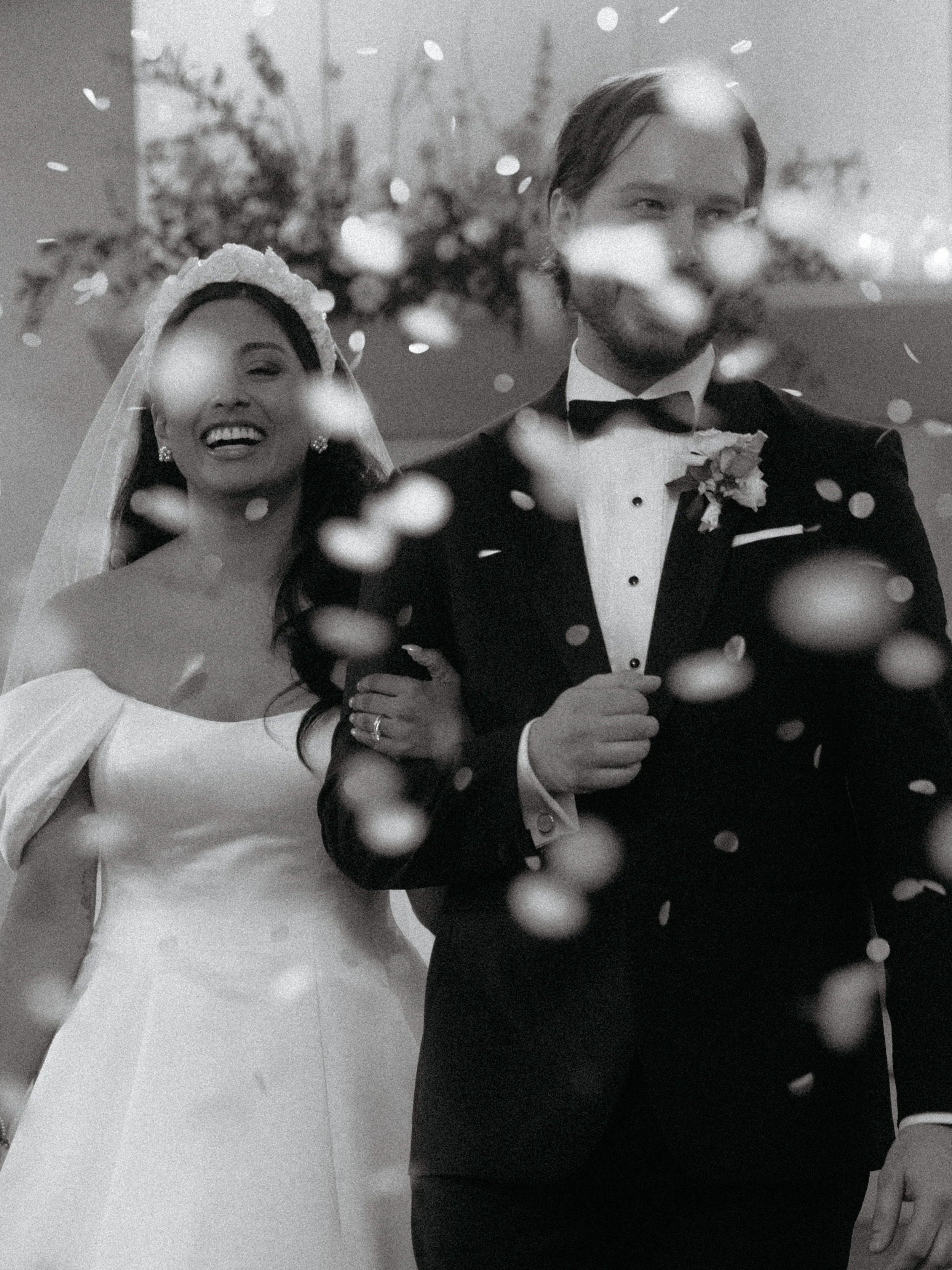 Black and white photo of a bride and groom at their wedding, with confetti falling around them. The bride is smiling, wearing a wedding gown and veil, and the groom is standing beside her in a tuxedo with a bow tie and boutonniere.