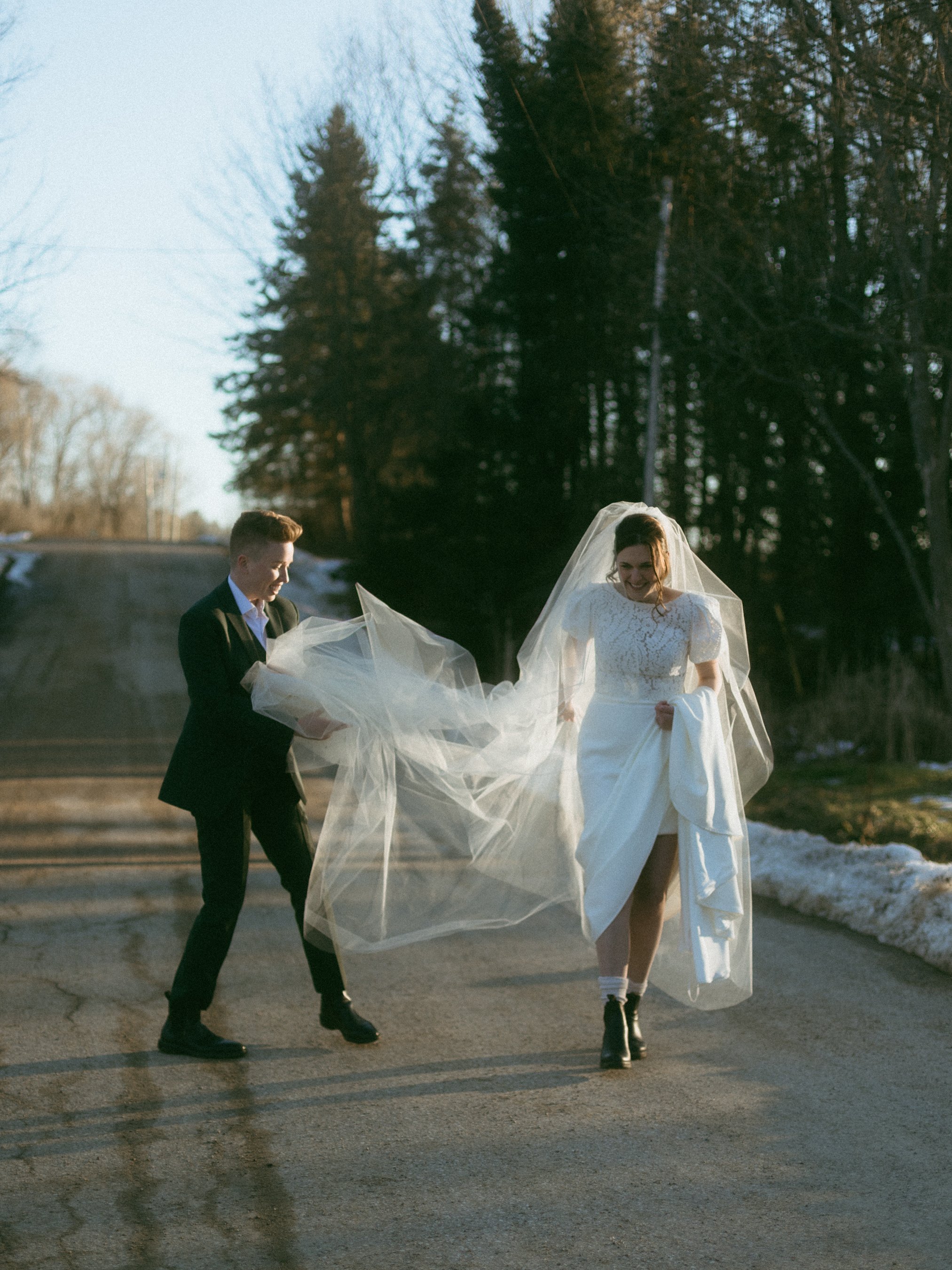 A bride and groom holding a wedding dress and veil outside on a sunny day, with trees and patches of snow in the background.