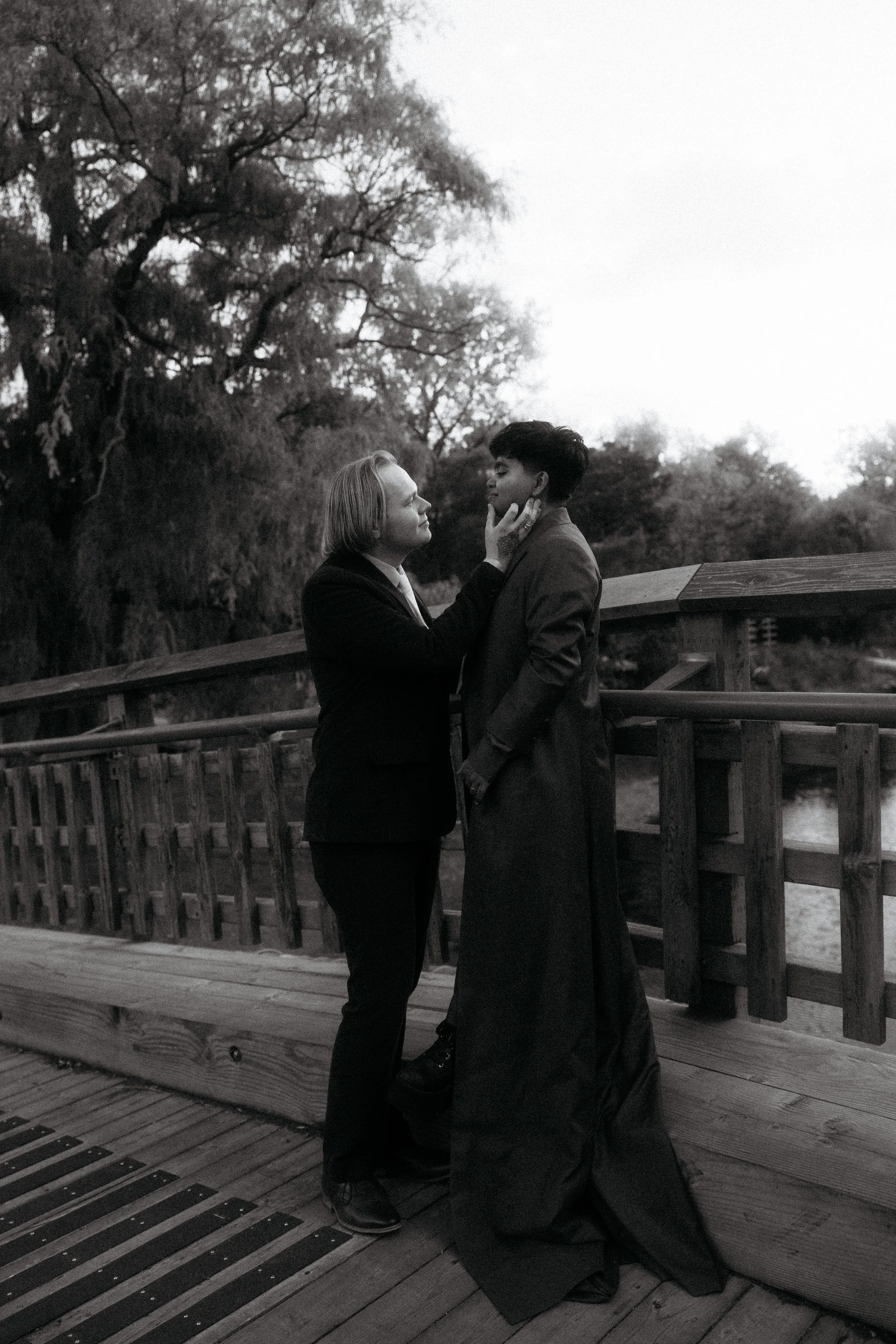 A woman and a person in a long dress share an intimate moment on a wooden bridge outdoors, with trees in the background, in black and white.