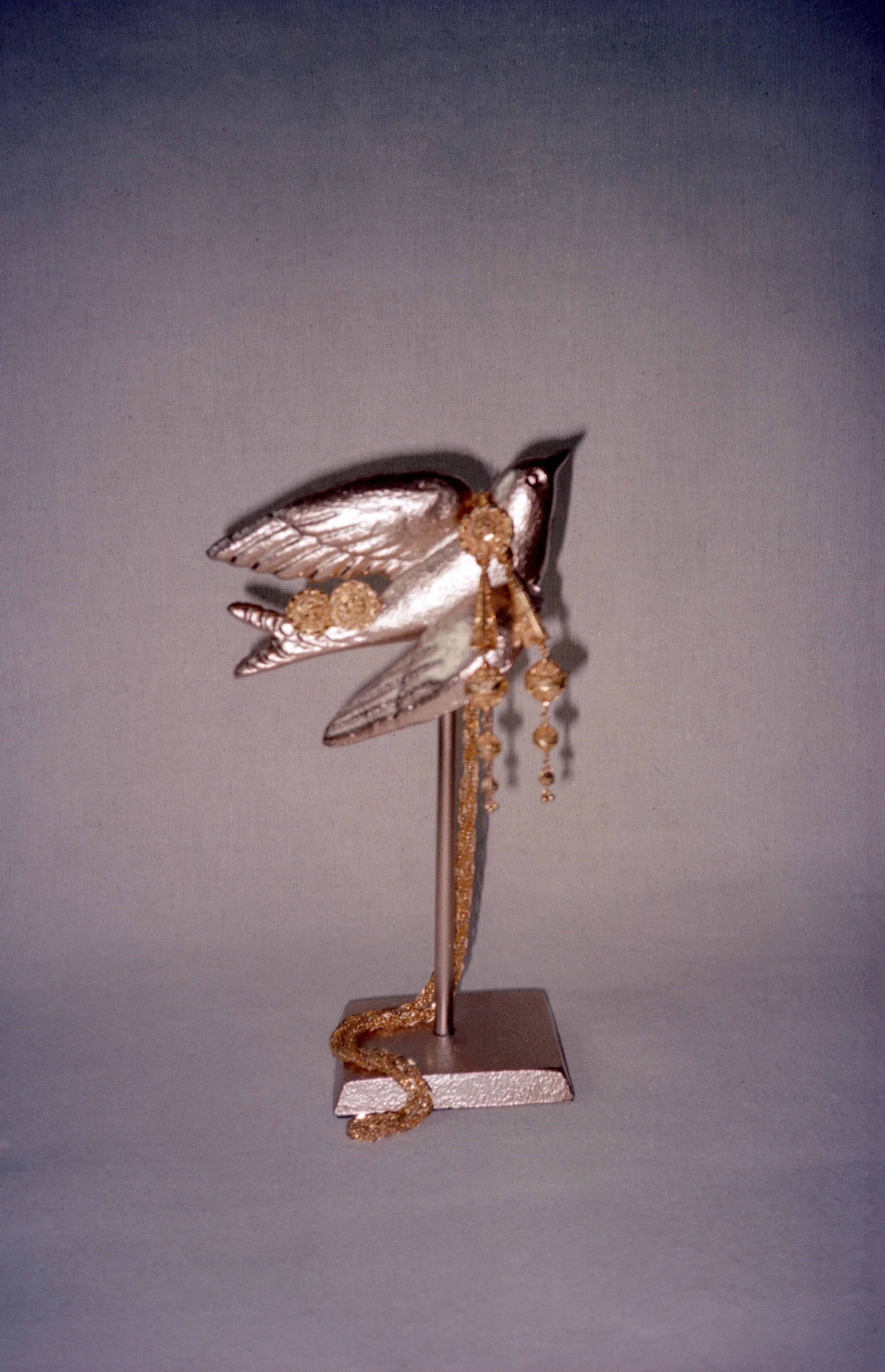 A metallic fairy sculpture with embellishments of gold beads and chains, mounted on a slender stand.
