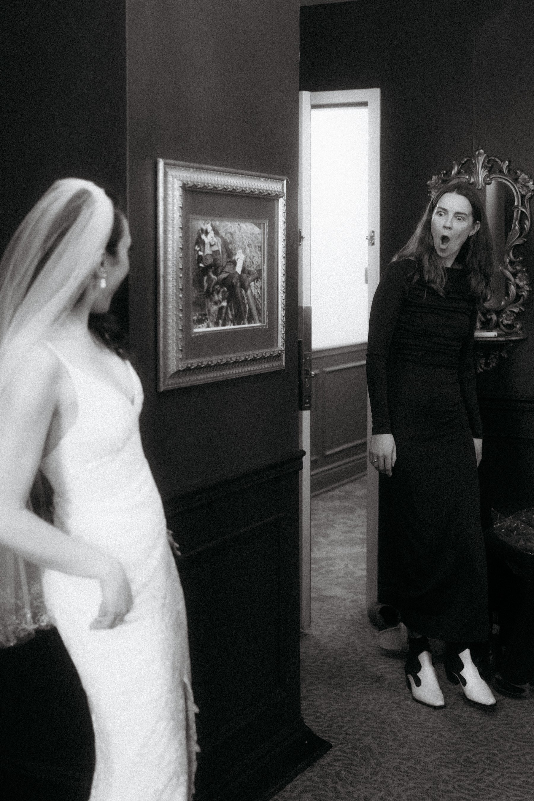 A woman in a black dress with white high-heeled boots is standing with her mouth open in surprise or shock, facing a woman dressed as a bride in a white wedding gown with a veil, inside a dimly lit room decorated with framed art and a decorative mirr