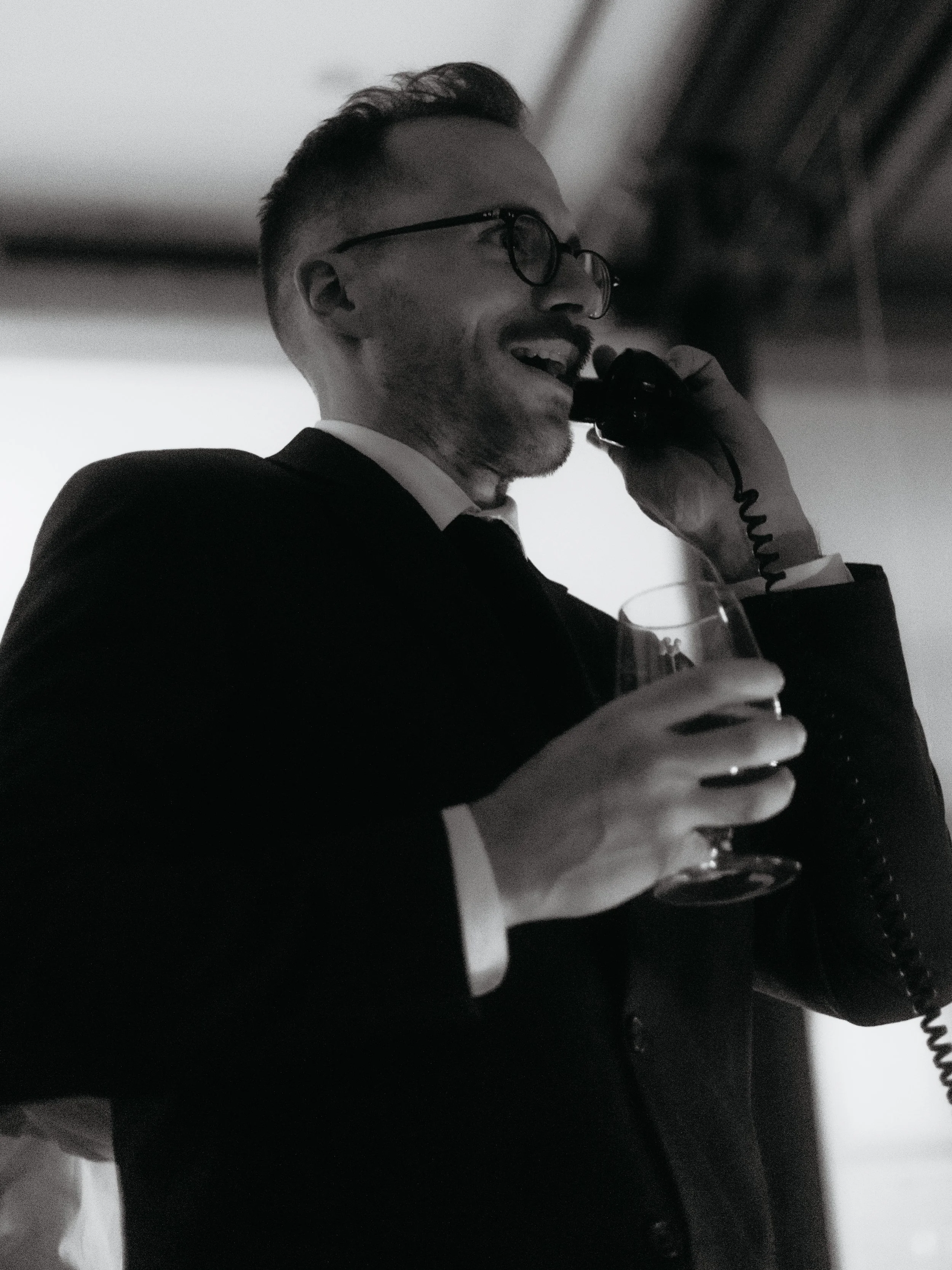Man in a suit and glasses holding a glass of wine and speaking on a corded phone.