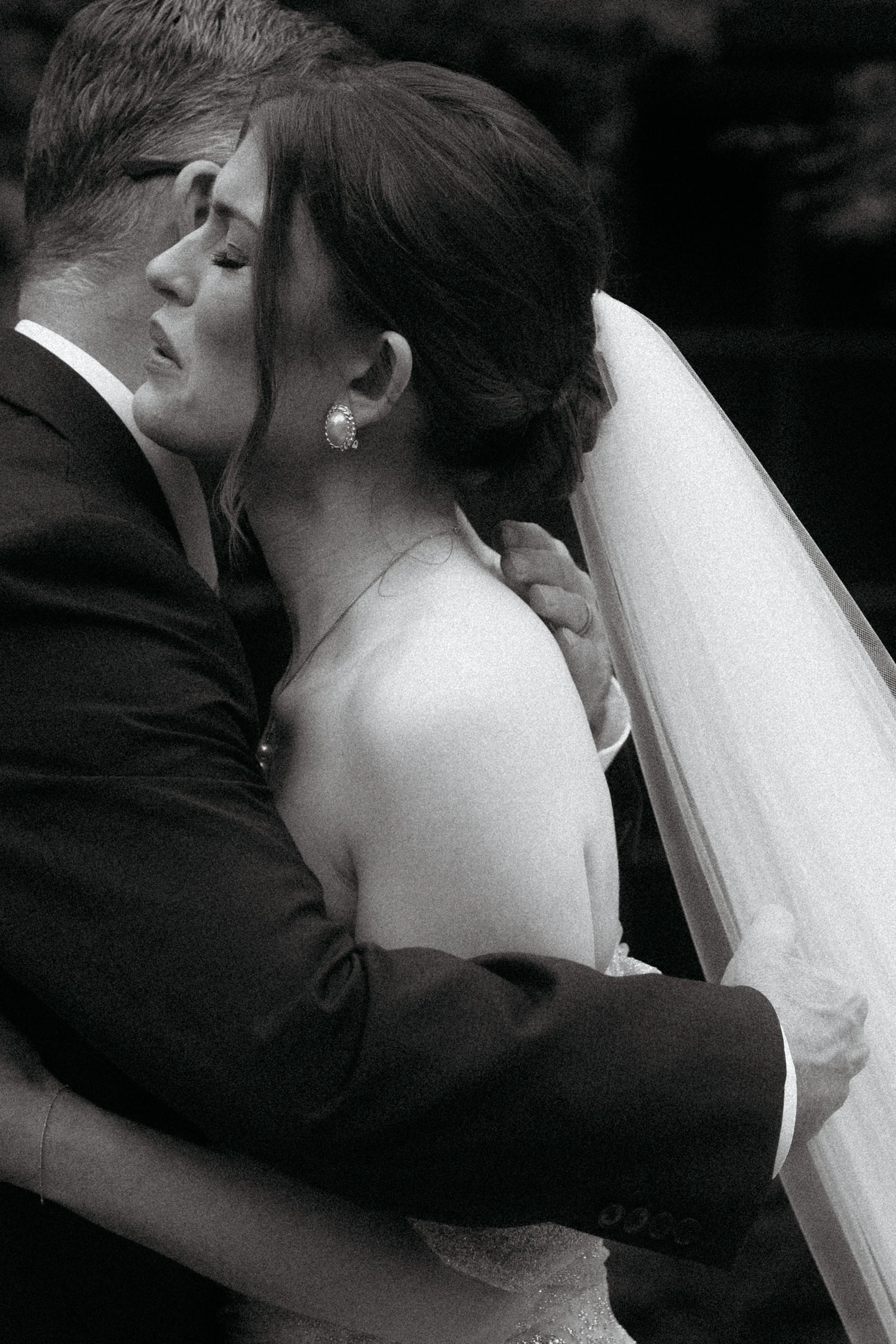 A black and white photo of a bride and groom sharing a kiss during their wedding ceremony, with the bride wearing earrings and a veil, and the groom in a suit.