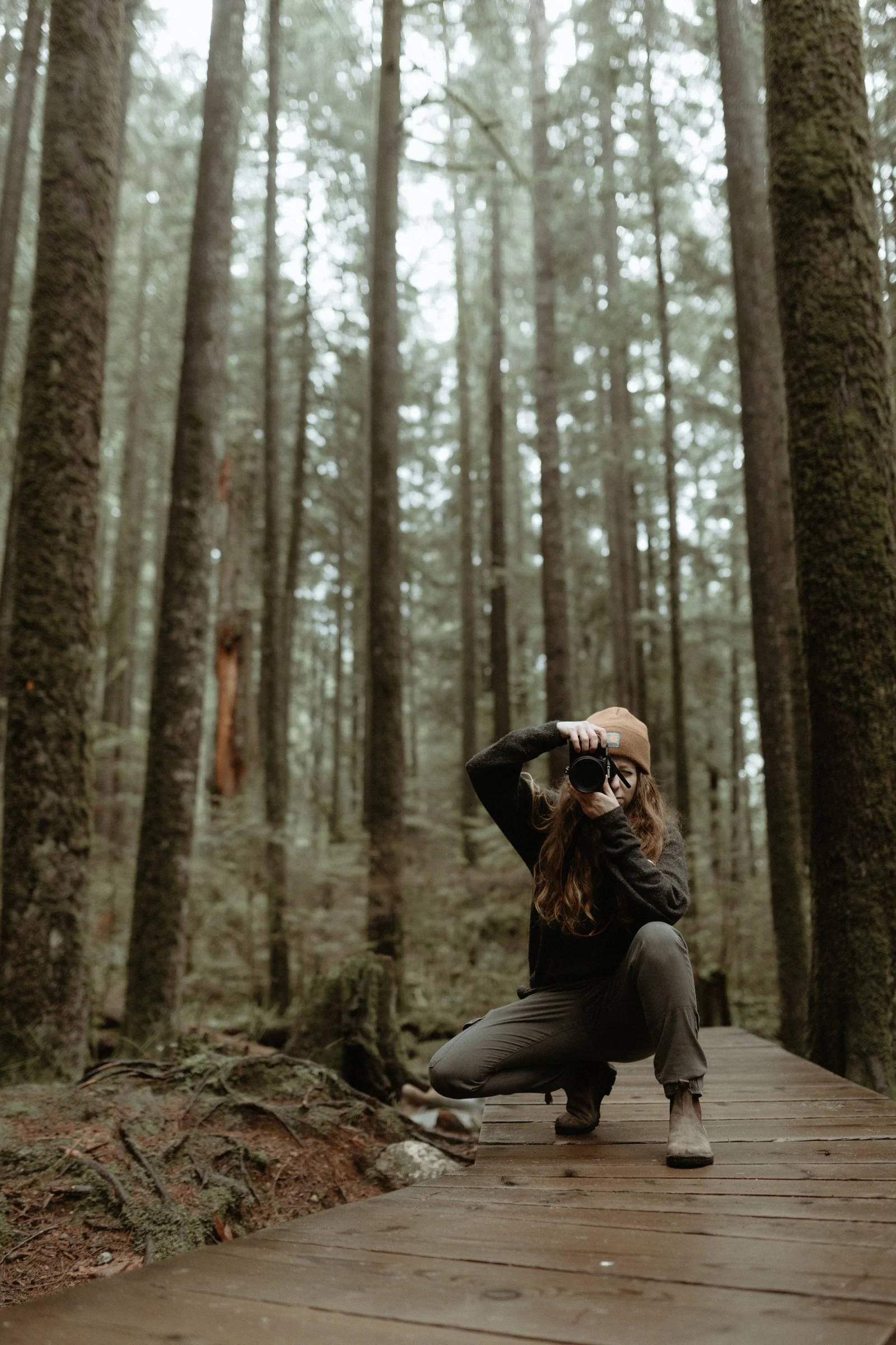 A woman crouching on a wooden boardwalk in a forest, holding a camera to her face, surrounded by tall trees with dense foliage.