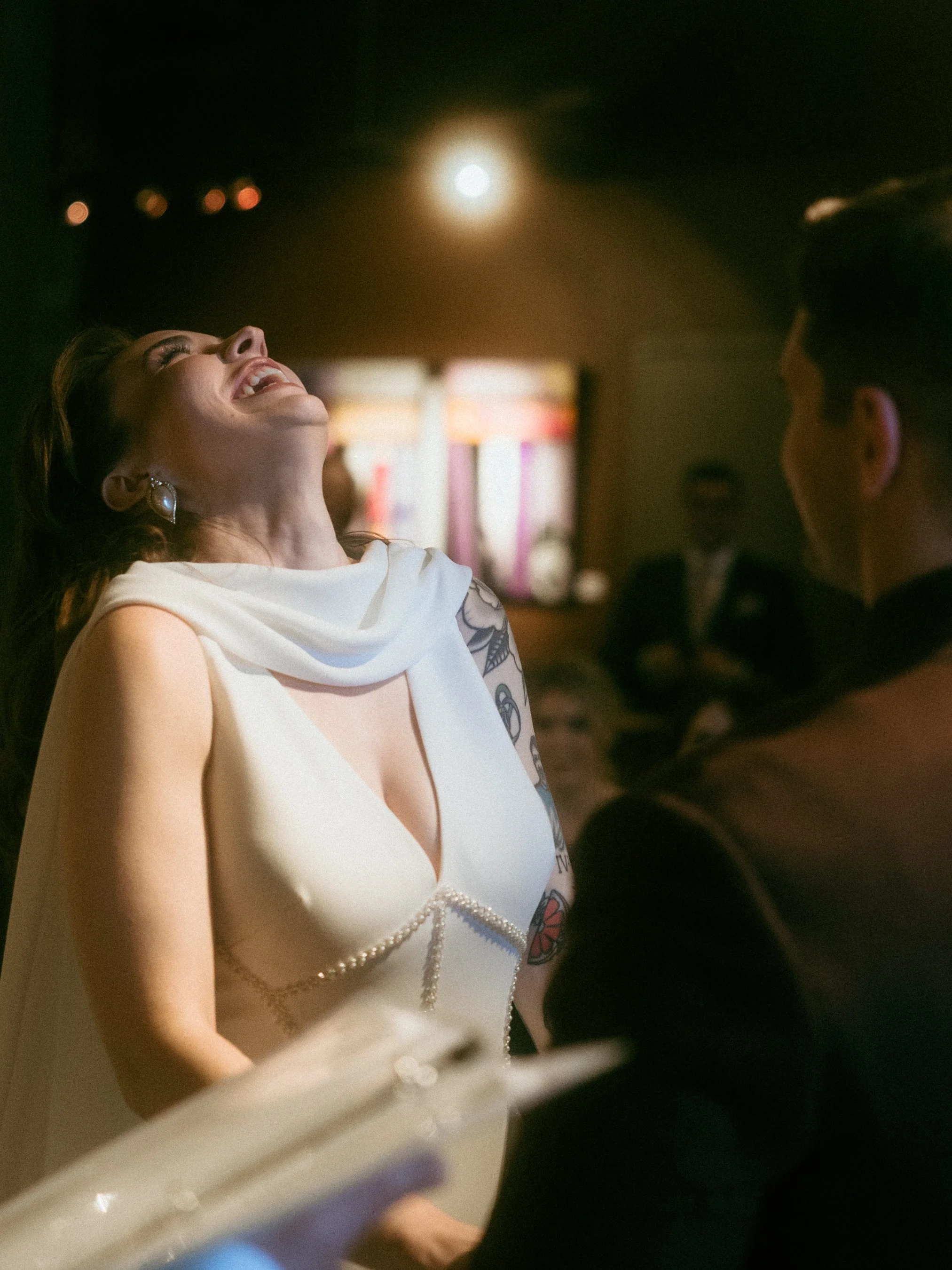 A woman with tattoos on her arm, wearing a white dress with a pearl detail, is laughing or smiling with her head tilted back. She is standing indoors at night, with a brightly lit area behind her and a full moon visible through a window or opening.