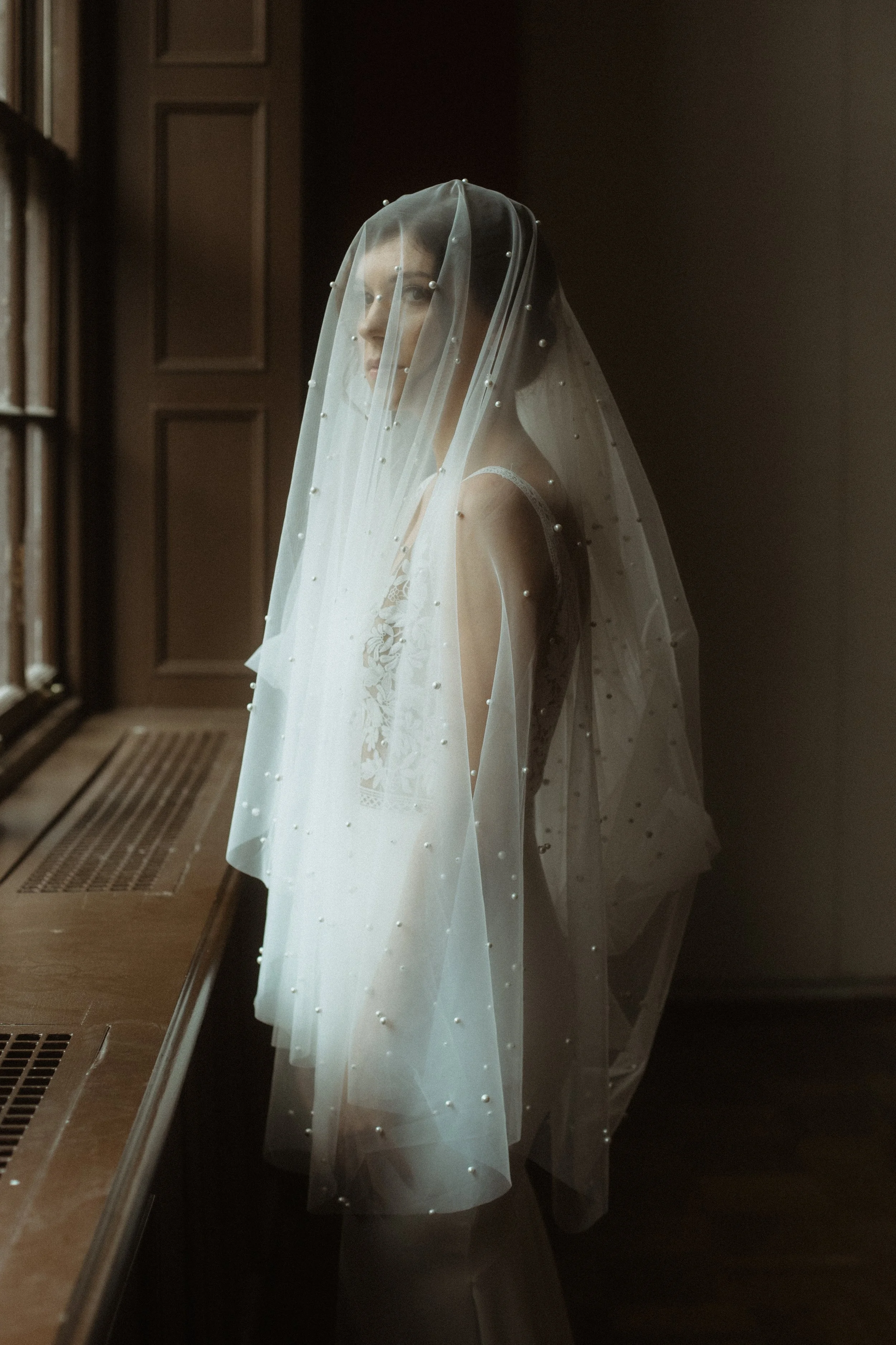 A bride in a lace wedding dress with a pearl-embellished veil looking out a window in a dimly lit room.