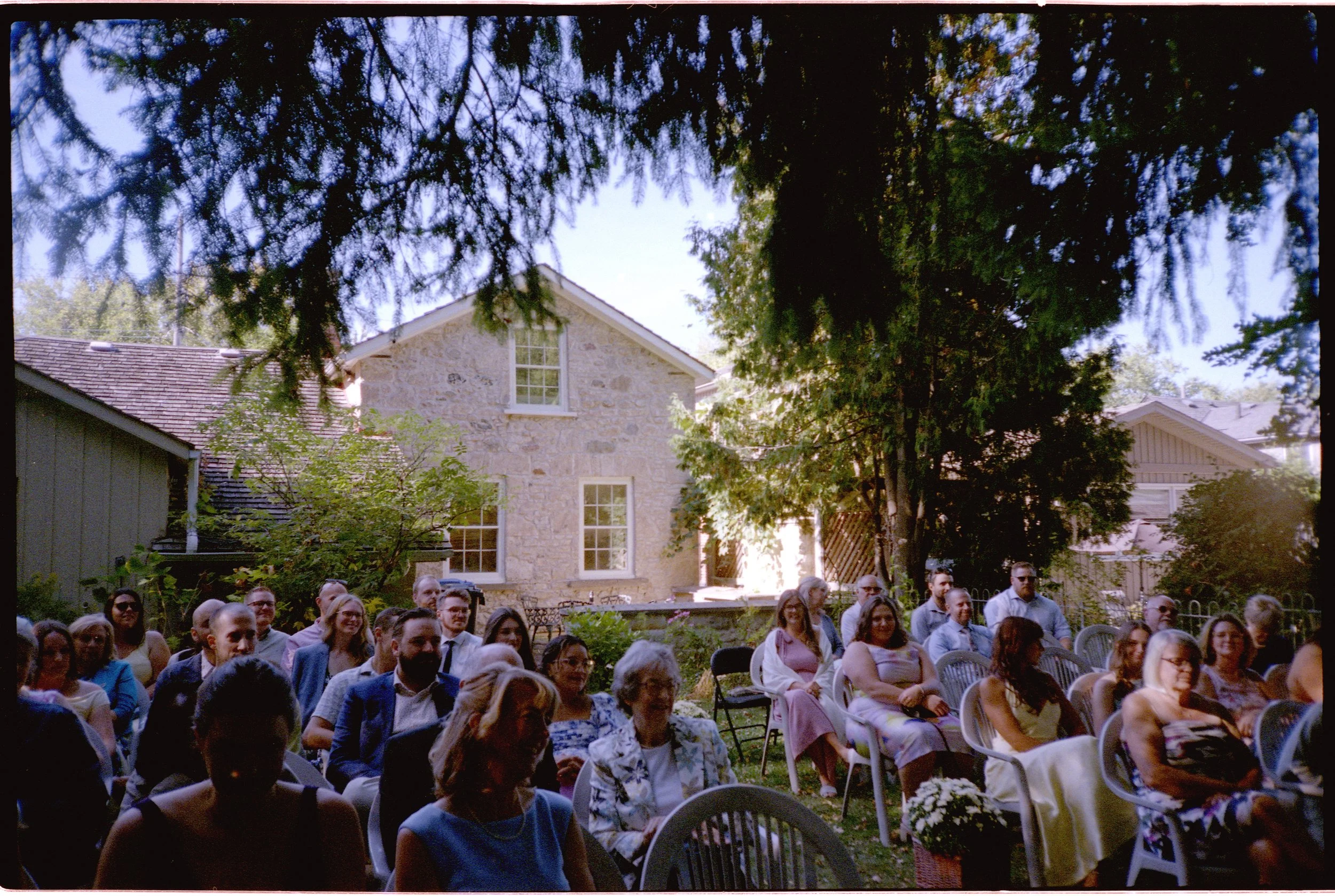 People seated outdoors during a gathering or event in a garden, with trees and a stone house in the background.