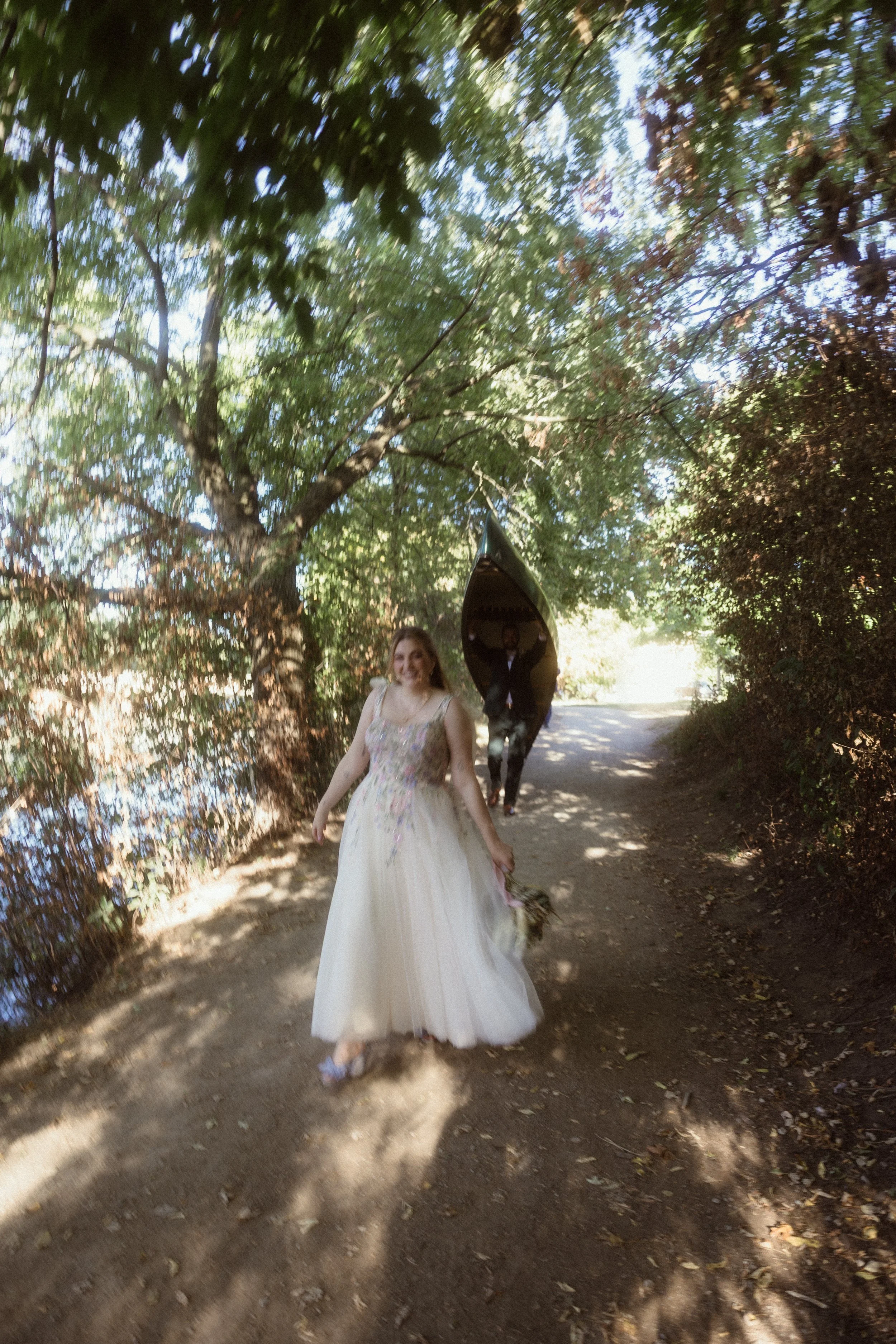 A woman in a white wedding dress walking on a shaded dirt path surrounded by trees, holding a bouquet, with a man in the background holding a canoe.