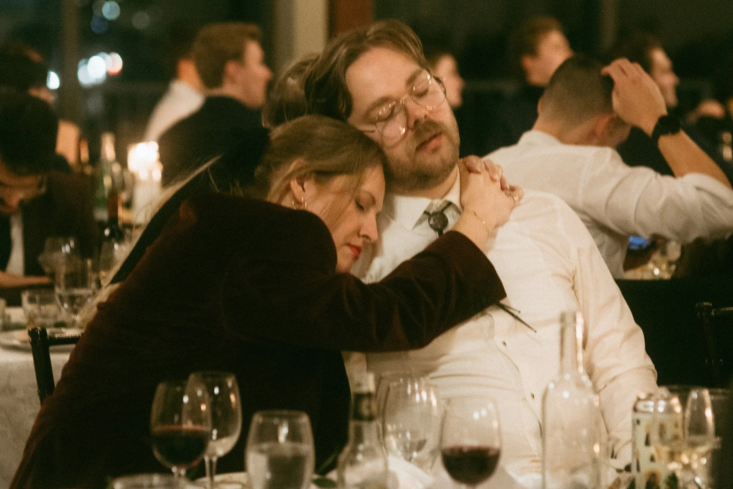 A woman and man are sitting closely with the woman resting her head on the man's shoulder and holding his neck, appearing to be asleep or resting at a social dinner event.