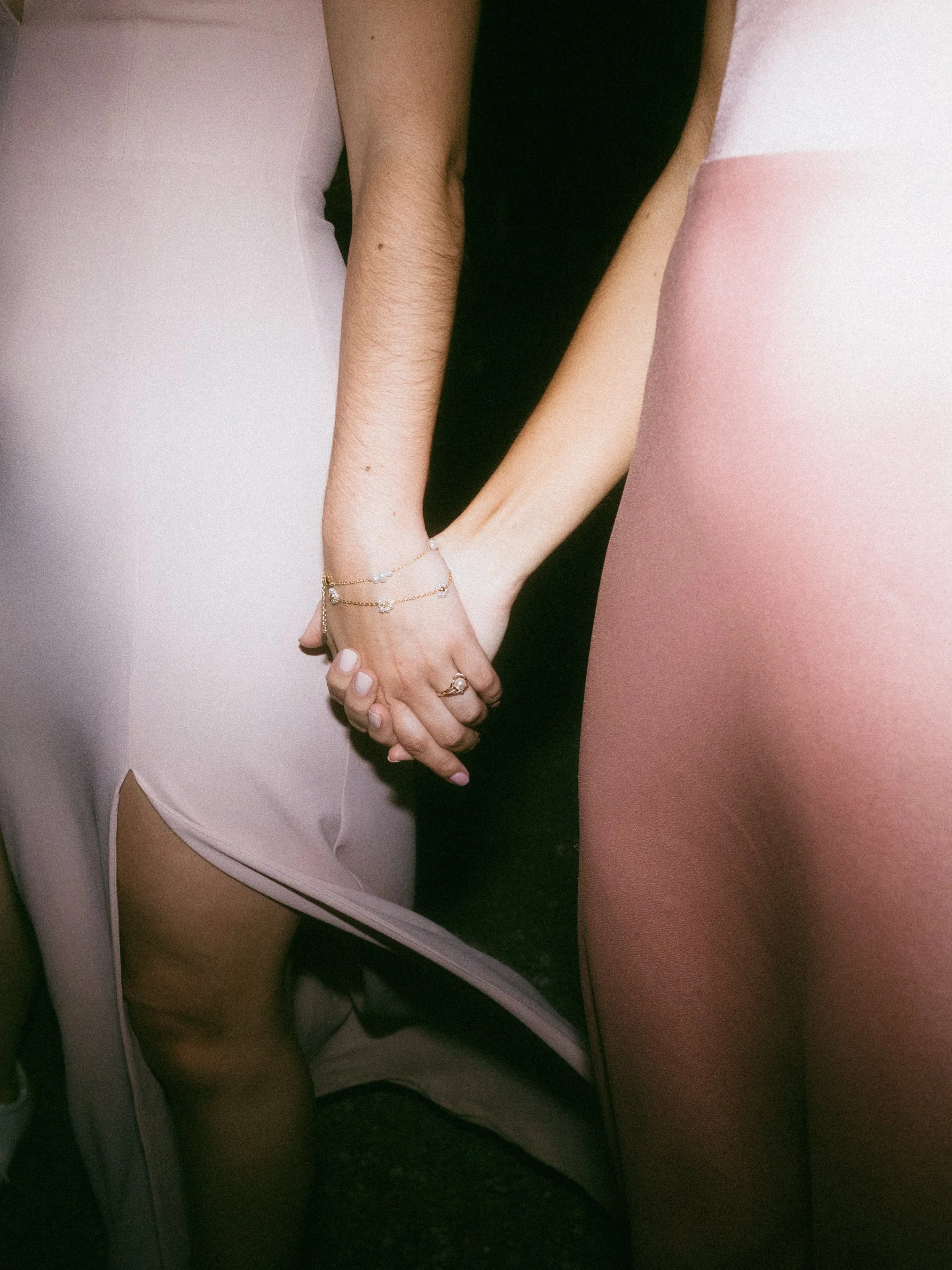 Close-up of two women holding hands, one wearing white dress with jewelry and the other in pink satin dress.