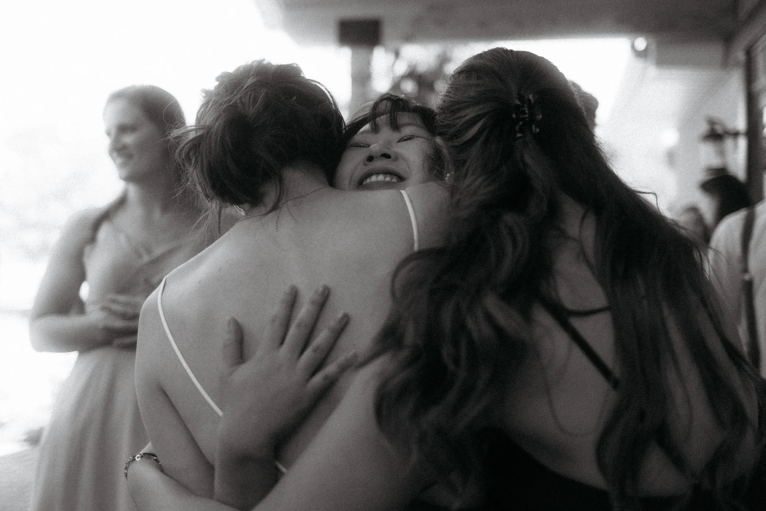 Three women hugging each other closely, smiling and appearing happy at a celebration or gathering, in a black and white photo.