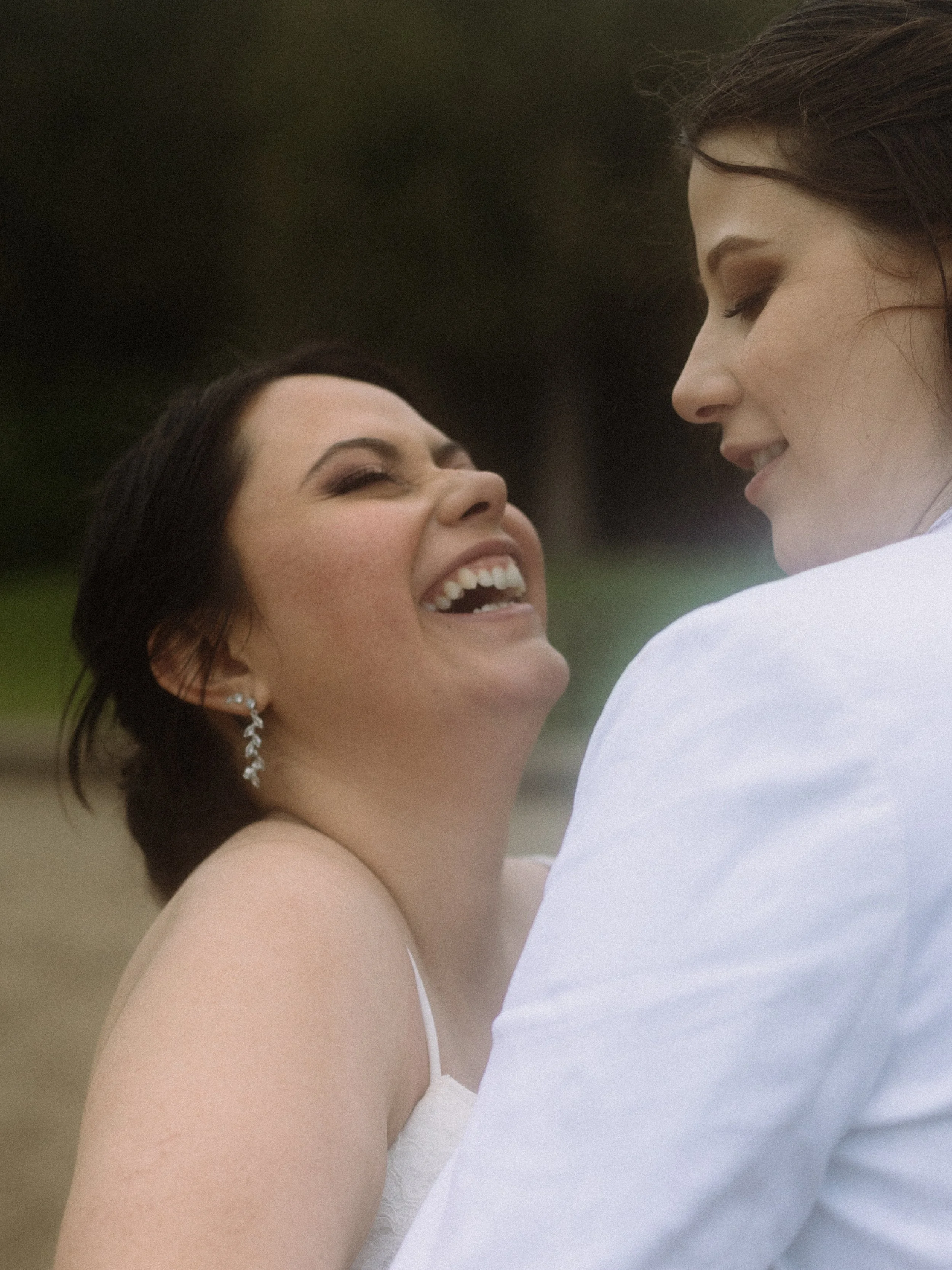 Two women sharing a joyful moment outdoors, one woman with dark hair and the other with brown hair, smiling and gazing at each other.