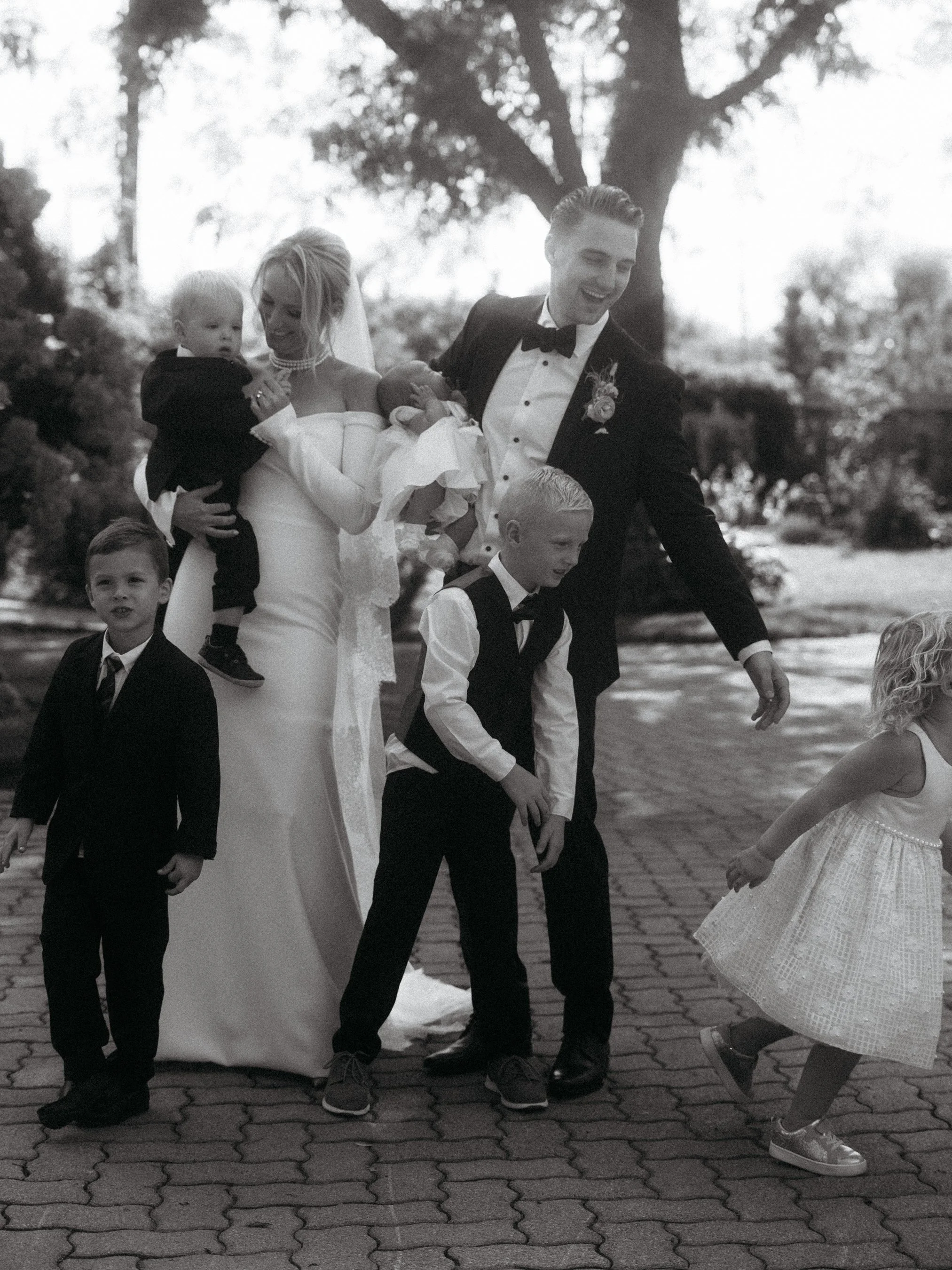 A black and white photo of a bride and groom with four children outdoors. The bride is holding a small child, and the groom is smiling with two other children nearby. All are dressed in formal attire, and trees are visible in the background.