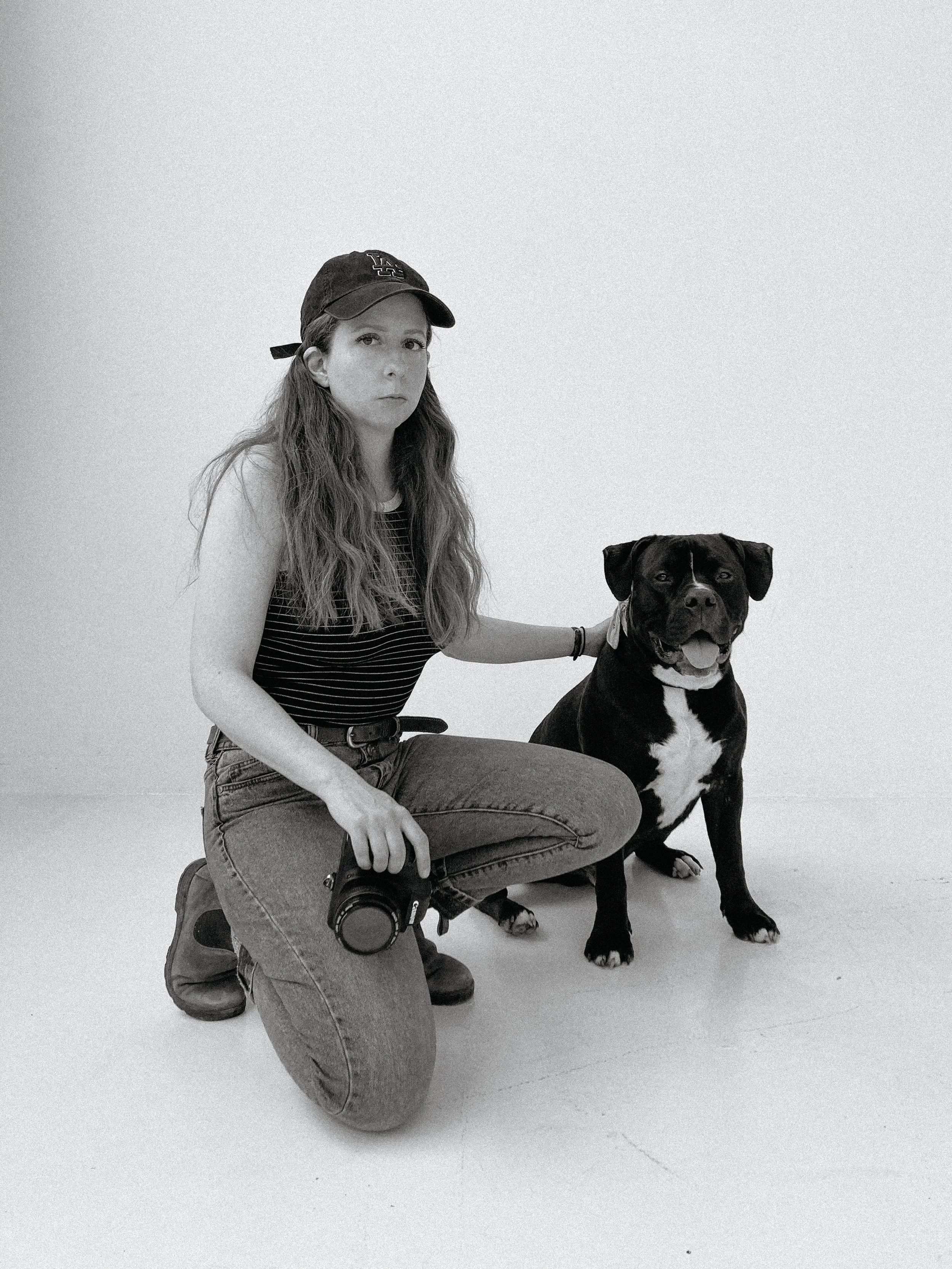 A young woman with long wavy hair, wearing a baseball cap, striped t-shirt, jeans, and boots, kneels on her right knee next to a large black and white dog, both looking at the camera in front of a plain white background.