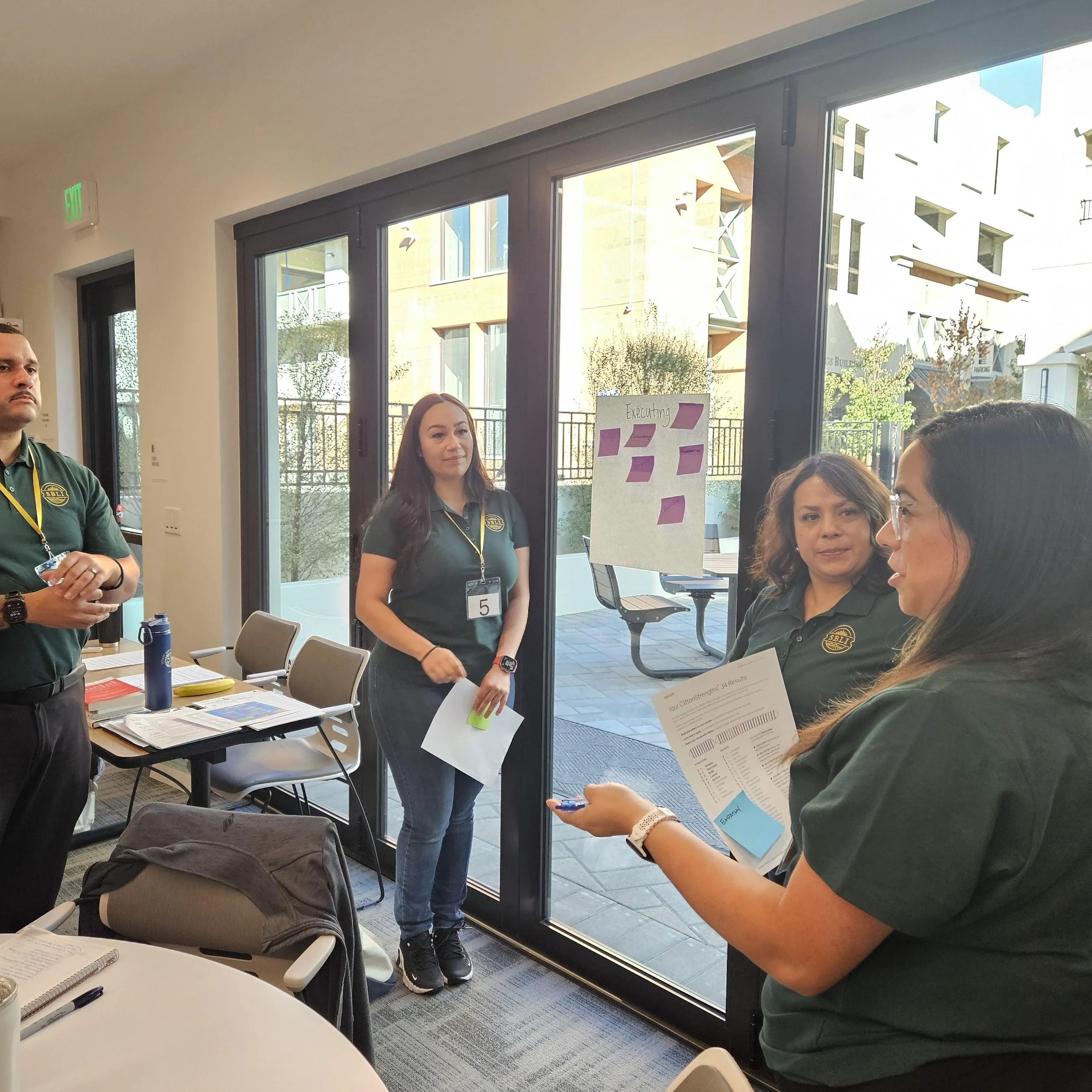 Four people in green shirts having a discussion near a conference table with papers and a water bottle, in a room with large glass doors showing a patio outside.