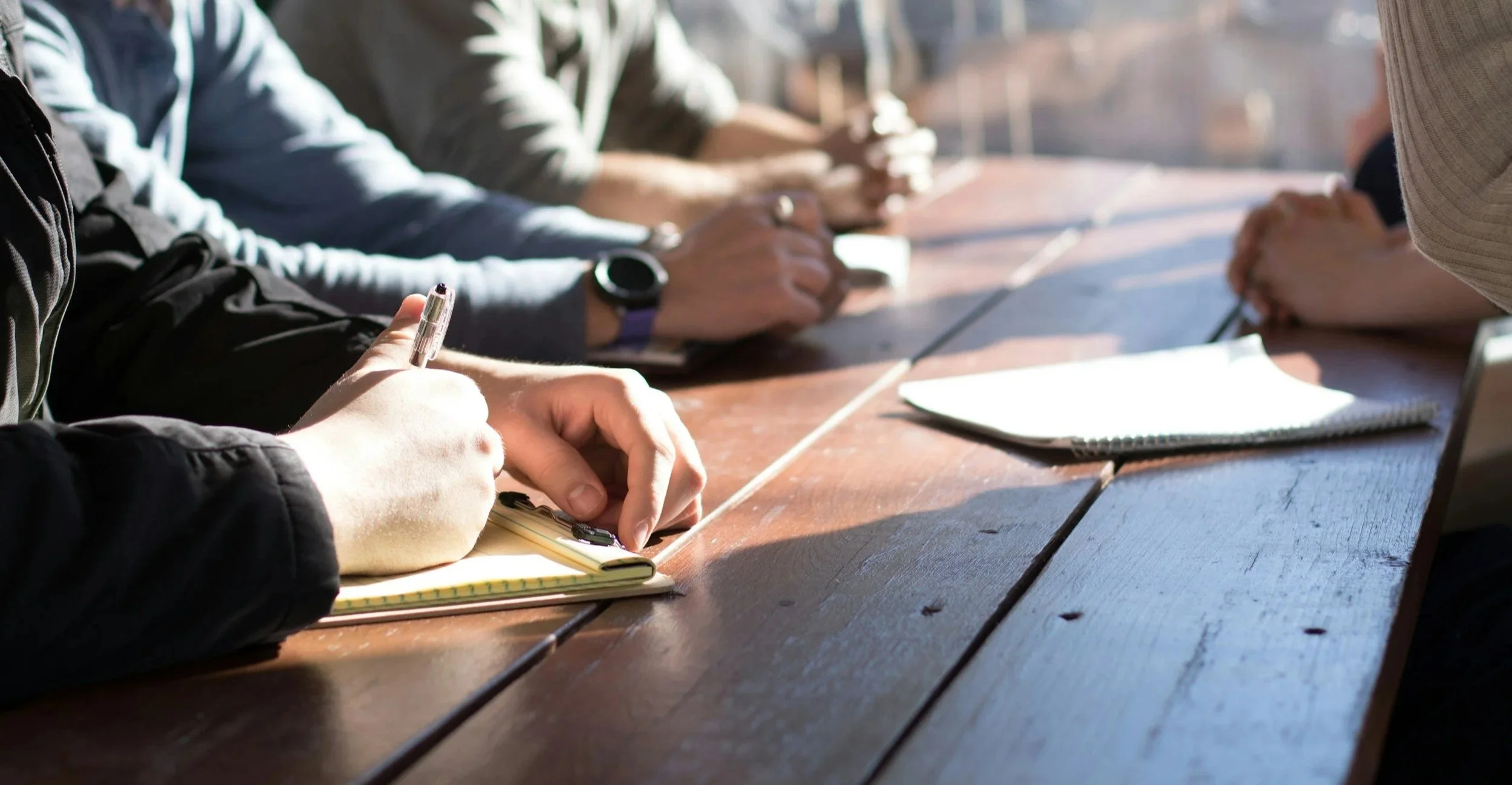 People sitting at a wooden table, taking notes and listening in a meeting or class, with notebooks and pens in front of them.