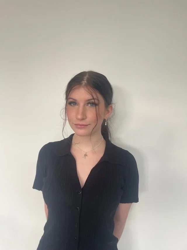 Young woman with dark hair wearing a black short-sleeved shirt, standing against a plain white wall.