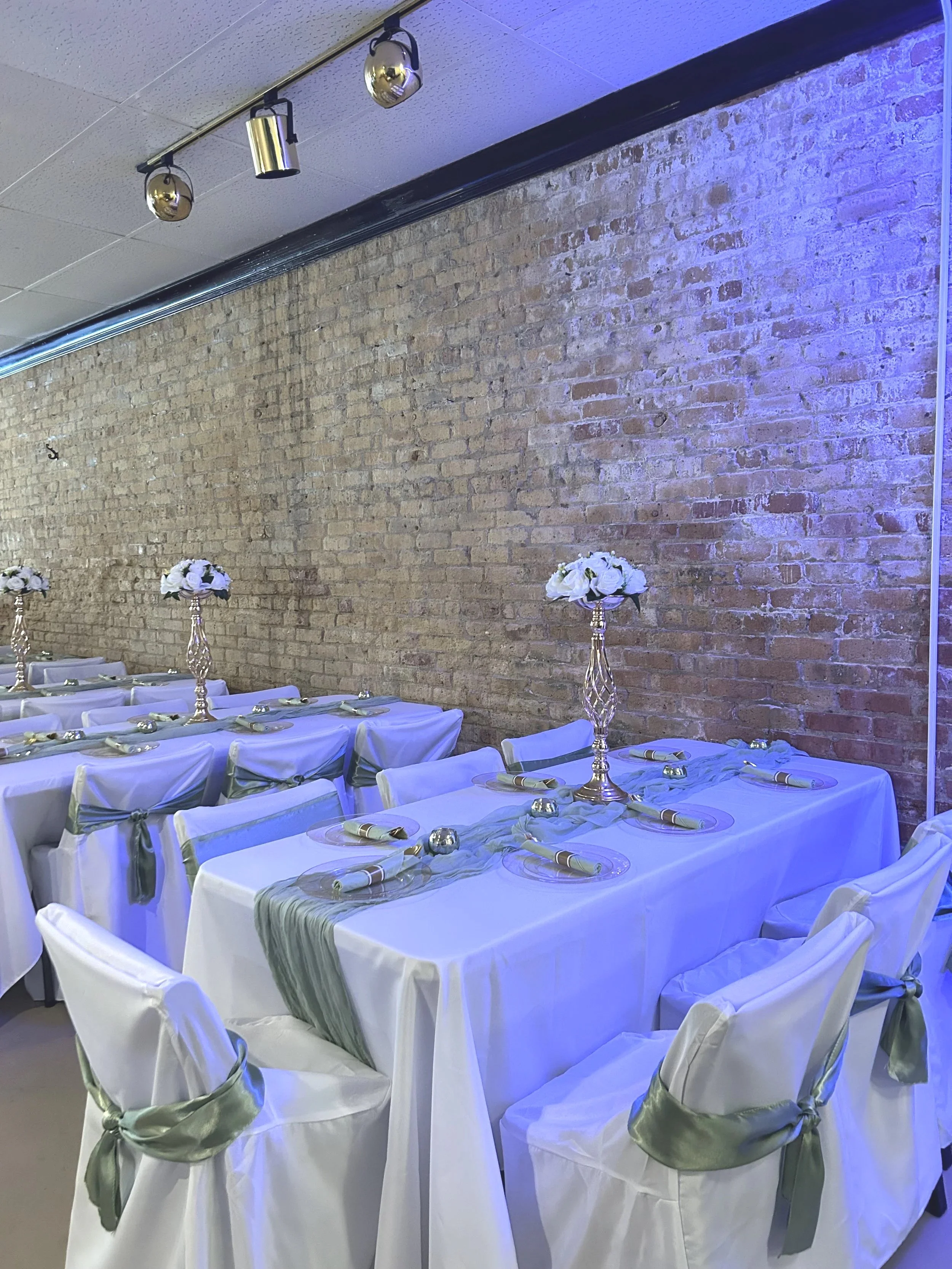 Decorated banquet table with white tablecloths, green sashes, and floral centerpieces in a room with an exposed brick wall and track lighting.