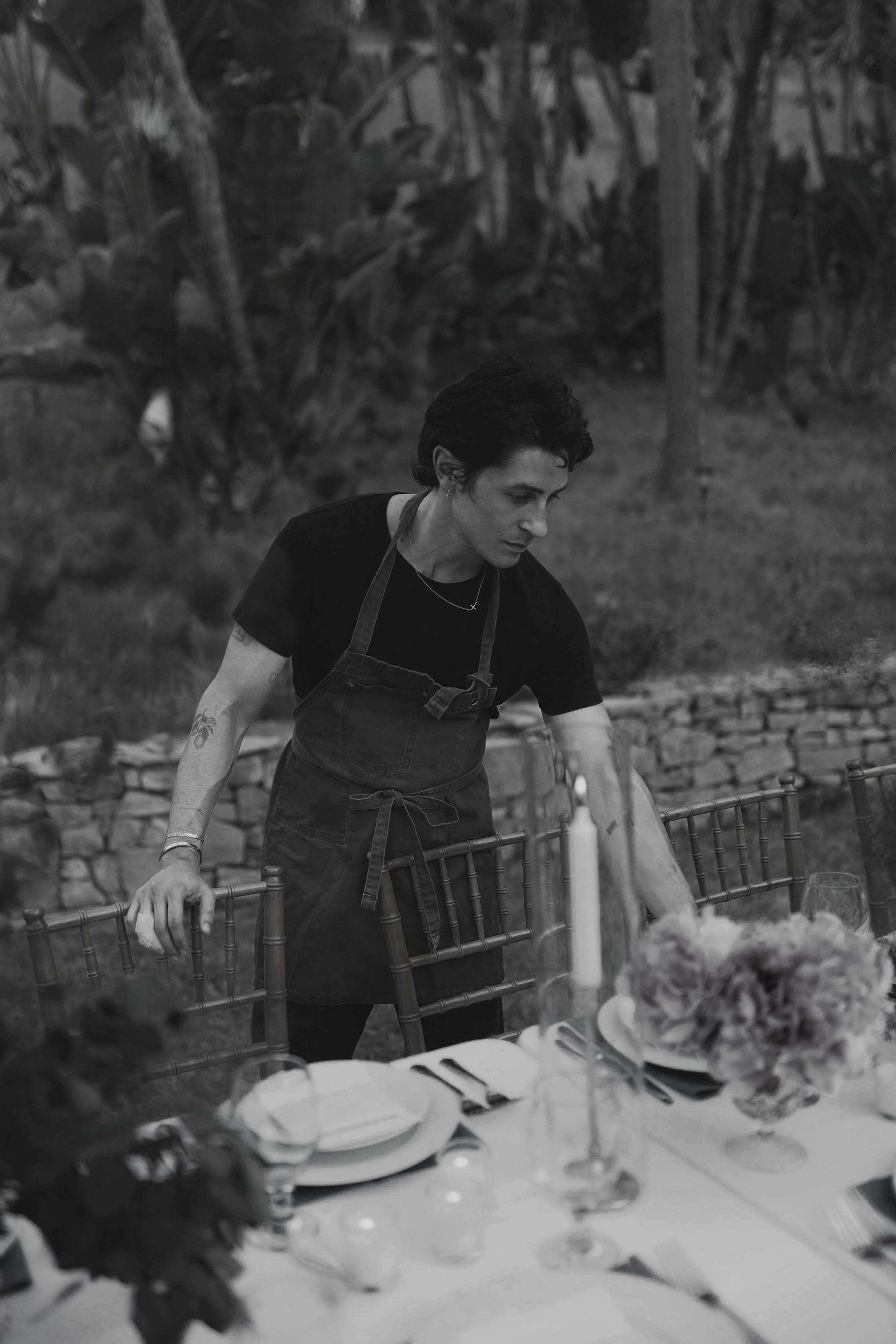 A woman with dark hair and tattoos wearing a black shirt and apron, setting a table outdoors.