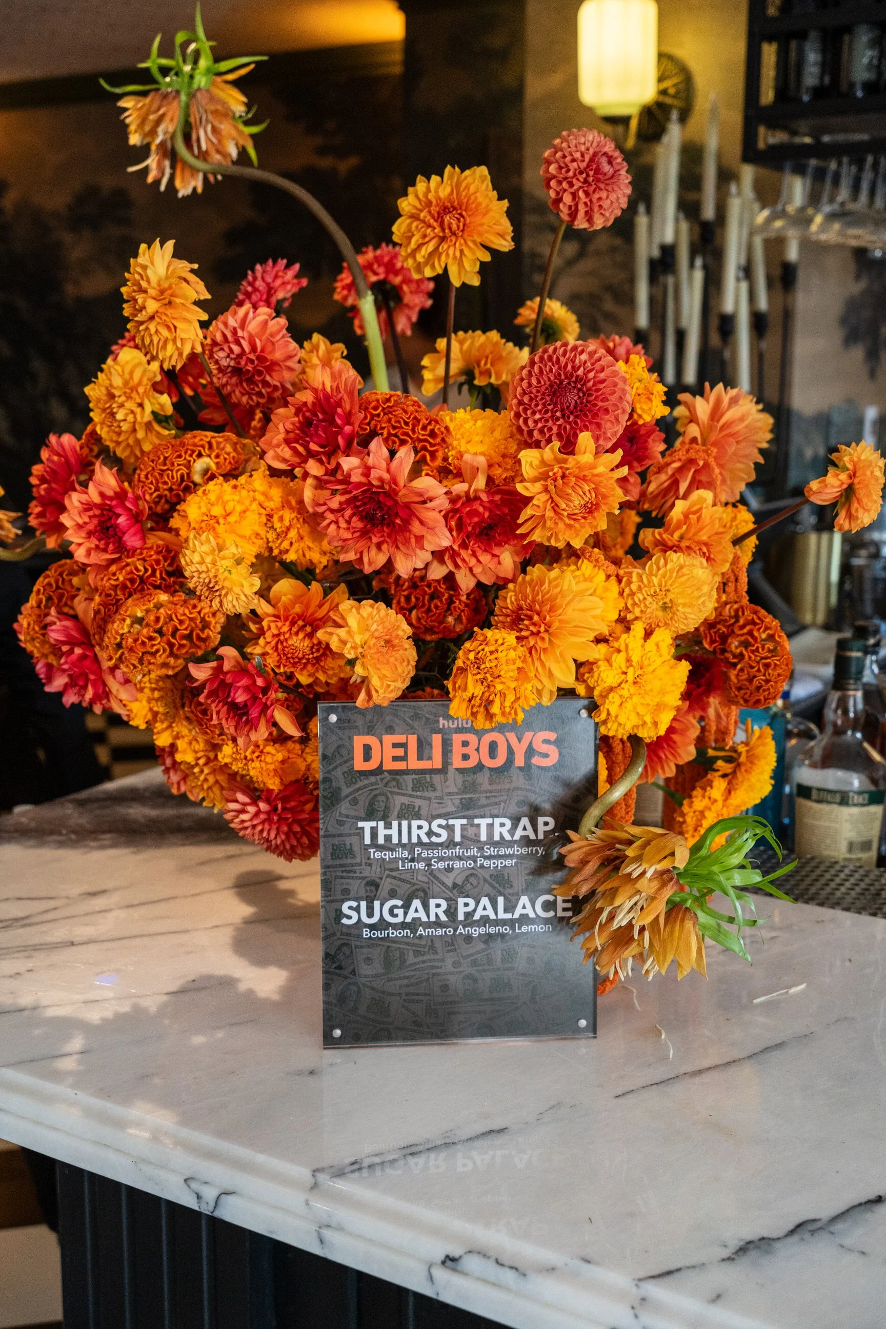 A flower arrangement with orange, pink, and yellow flowers behind a menu sign. The sign lists drinks: Deli Boys menu with Thirst Trap (Tequila, Passionfruit, Strawberry, Lime, Serrano Pepper) and Sugar Palace (Bourbon, Amaro Angeleno, Lemon). The bac
