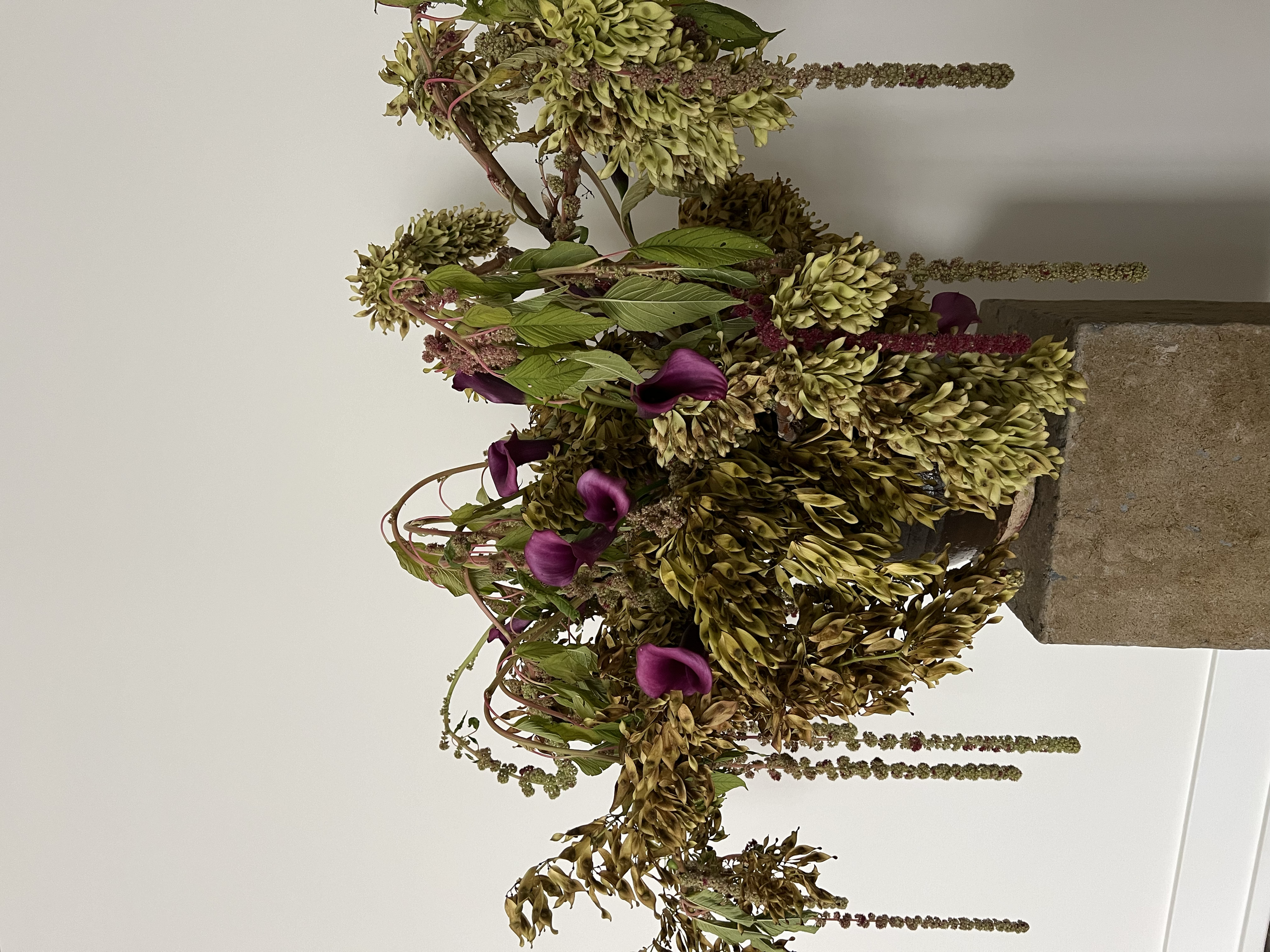 A floral arrangement with large clusters of greenish-yellow flowers, green leaves, and purple flowers, set against a plain white background. The arrangement is mounted on a light-colored textured base.