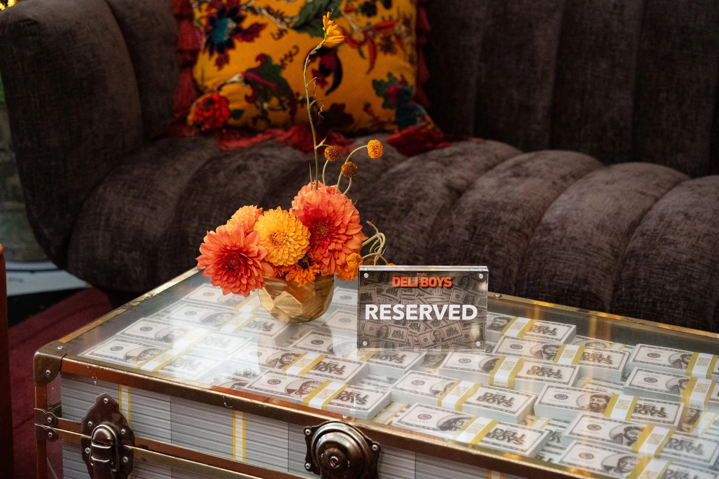 A coffee table covered with display stacks of dollar bills, a decorative bouquet of orange and yellow flowers, and a small sign that reads 'RESERVED.' In the background, there is a dark-colored sofa with throw pillows, including a bright yellow one w