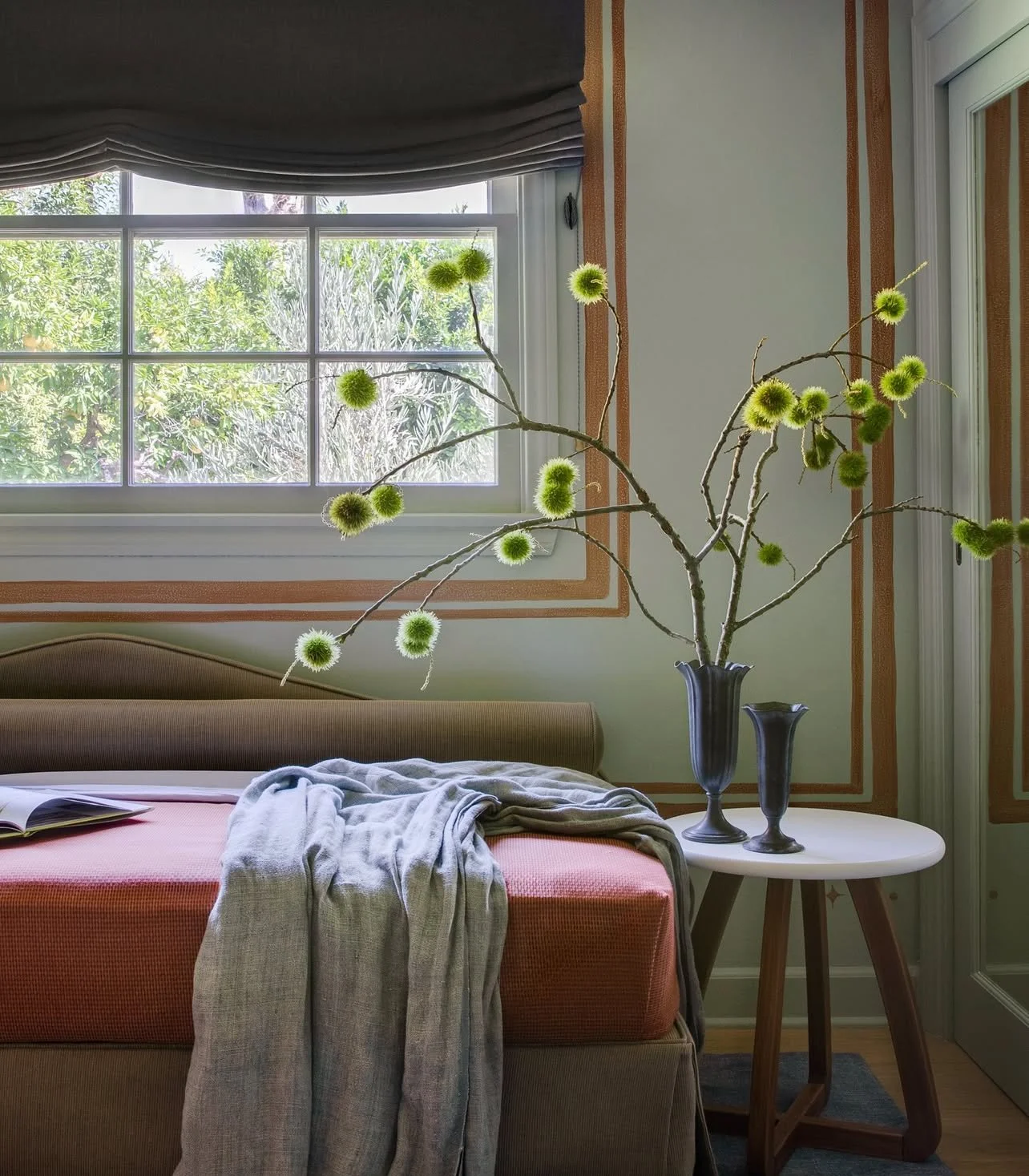 A bedroom with a window showing trees outside. Inside, there is a bed with a pink mattress, a light-colored blanket, slippers, and an open book. On a small white side table, there is a dark vase with branch-like stems and bright green fluffy flowers.