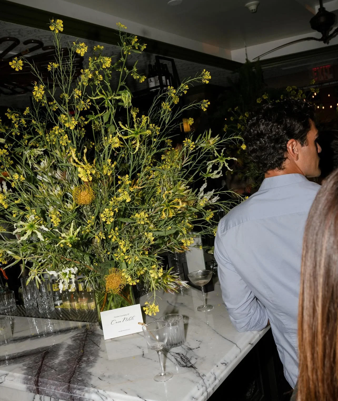 A large yellow and green floral arrangement on a marble table at a social event, with a man in a white shirt standing near the table and glasses of drinks around.