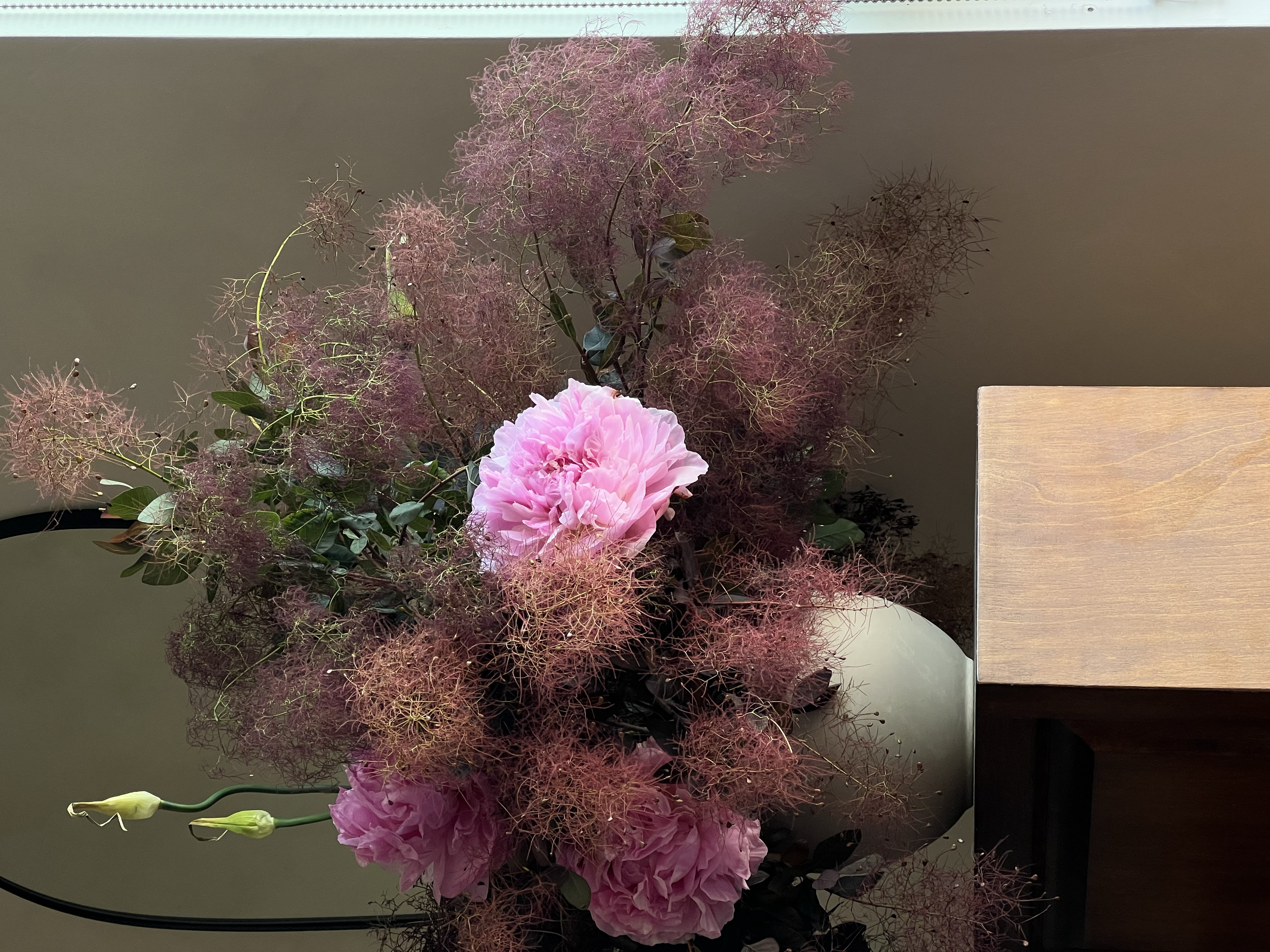 Arrangement of pink peony flowers, fluffy pink blooms, and wispy red and pink foliage on a table near a wall.