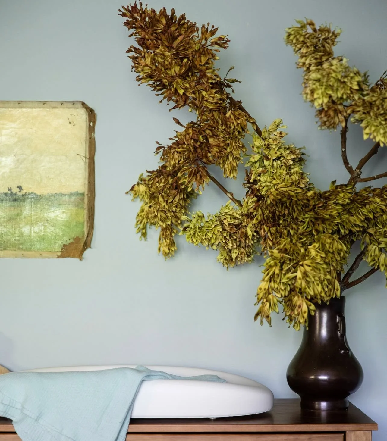 A large dark vase with dried yellow and brown flowers on a wooden table beside a bed with light blue bedding. A small painting hangs on the light blue wall.