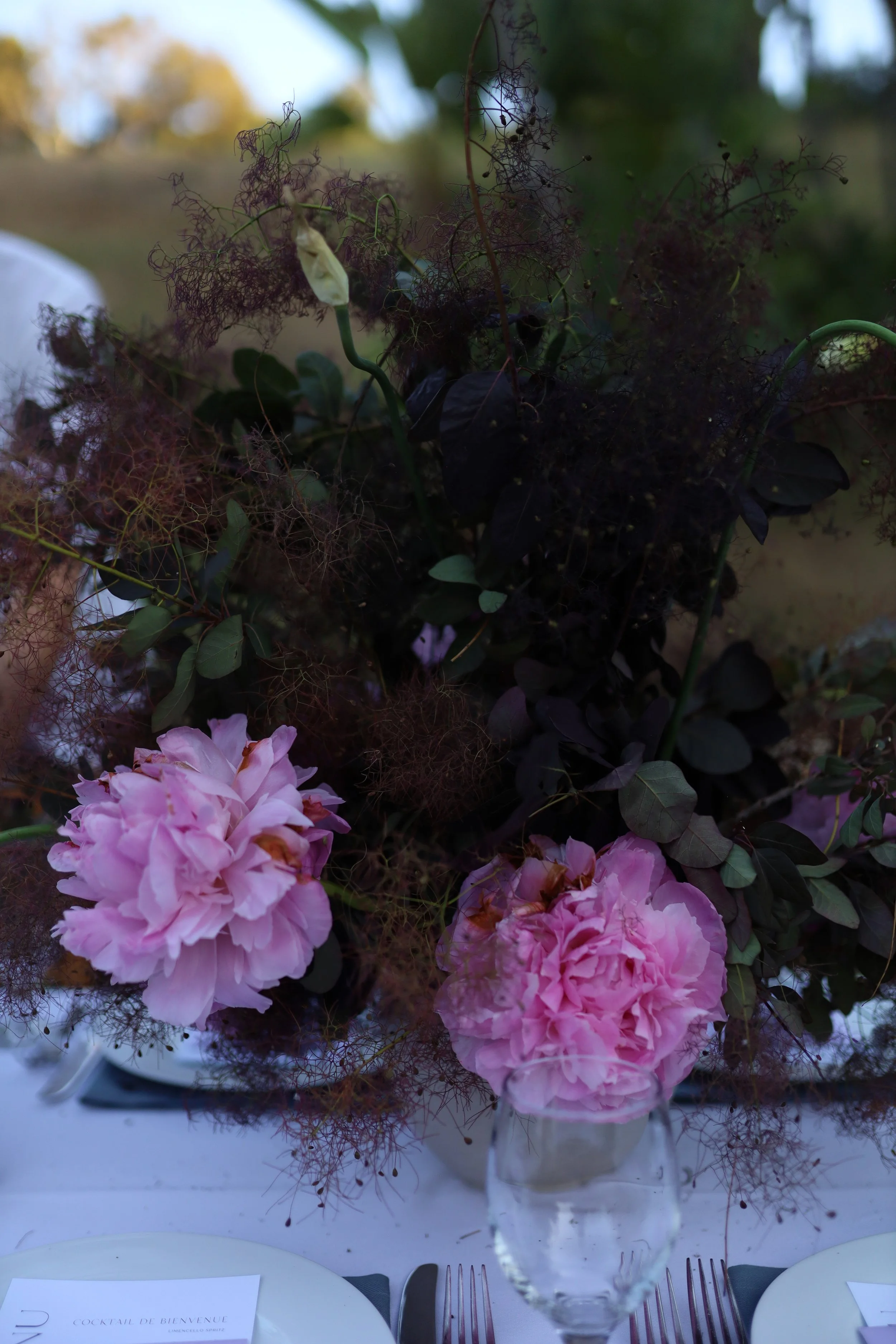 A floral arrangement in a glass vase with pink peonies and dark greenery, set on a table with white plates, silverware, and a welcome card, outdoors with blurred trees in the background.