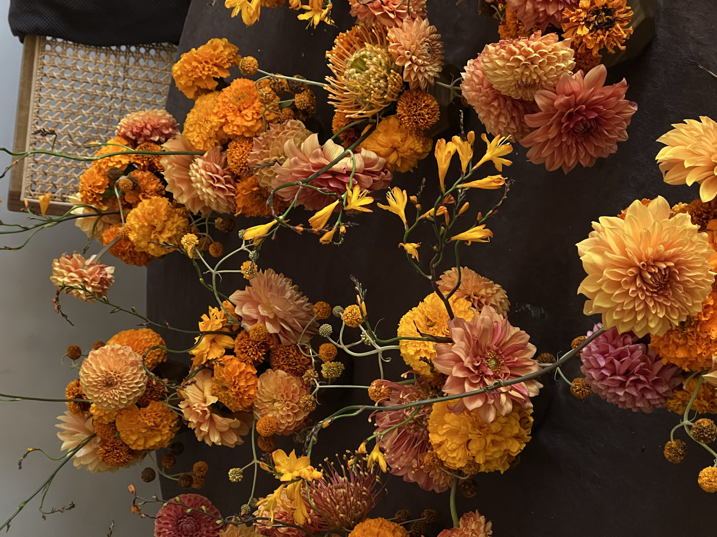 An assortment of orange, yellow, and peach-colored flowers, including dahlias, marigolds, and other blooms, with some long, green stems, laid out on a black surface.