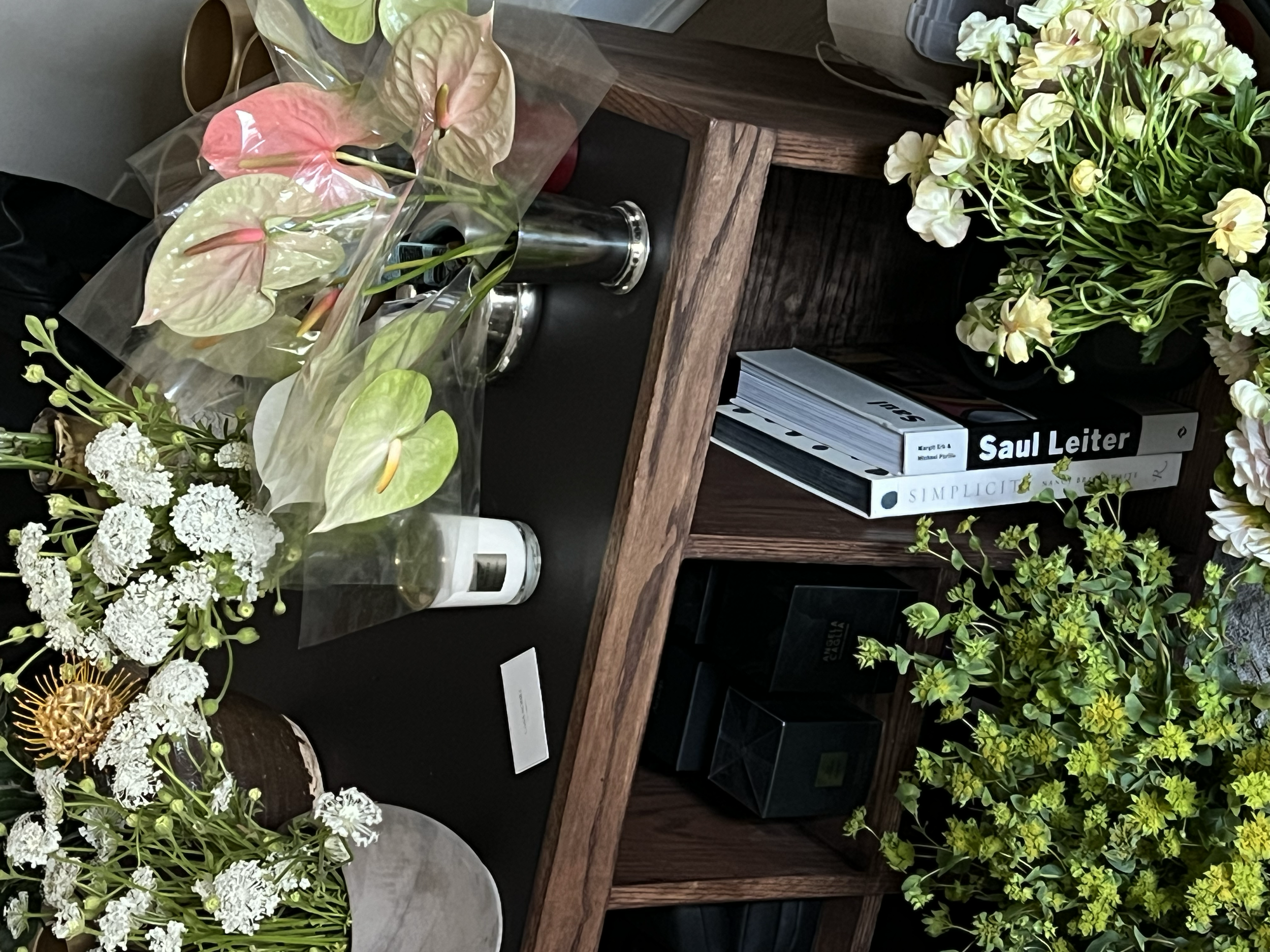 Various flowers, including white, pink, and yellow blooms, arranged on a wooden table and in containers.