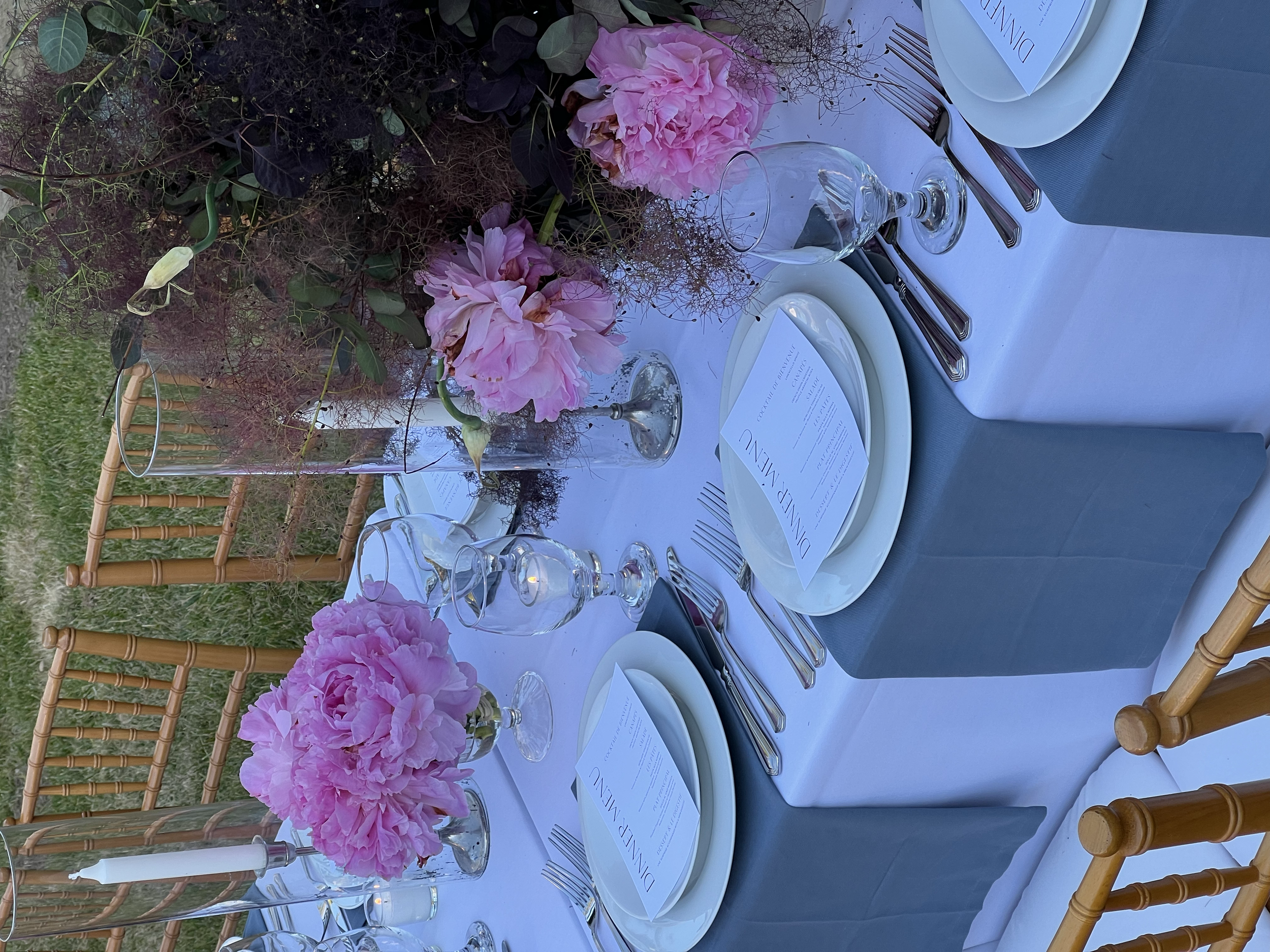A table setting with pink peony flowers in glass vases, white plates with printed menus, silverware, wine glasses, and a blue cloth napkin on a white tablecloth. A wooden chair is partially visible in the background.