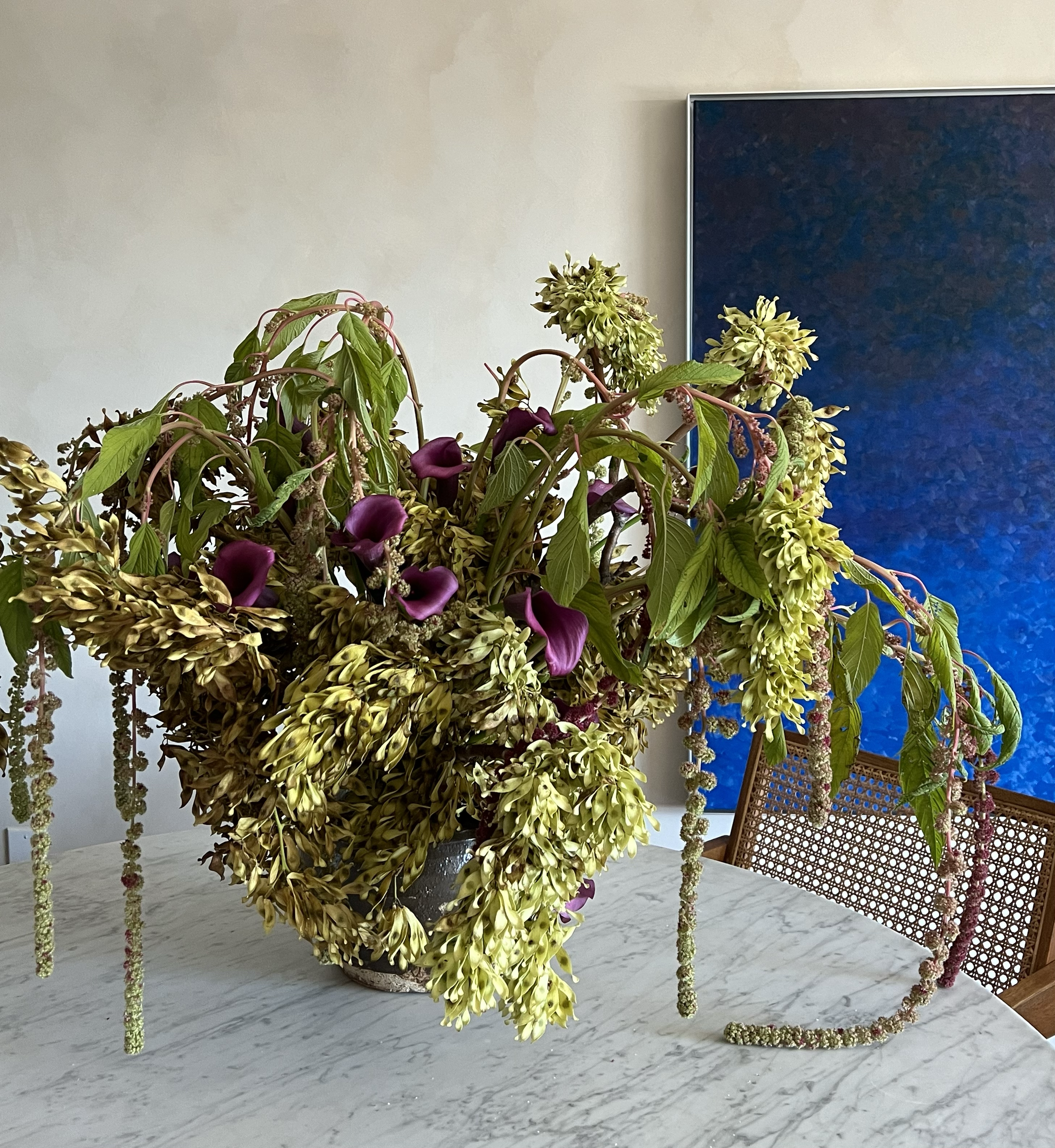 Flower arrangement with yellow, purple, and green flowers on a white marble table, with a modern chair and dark abstract art on the wall in the background.