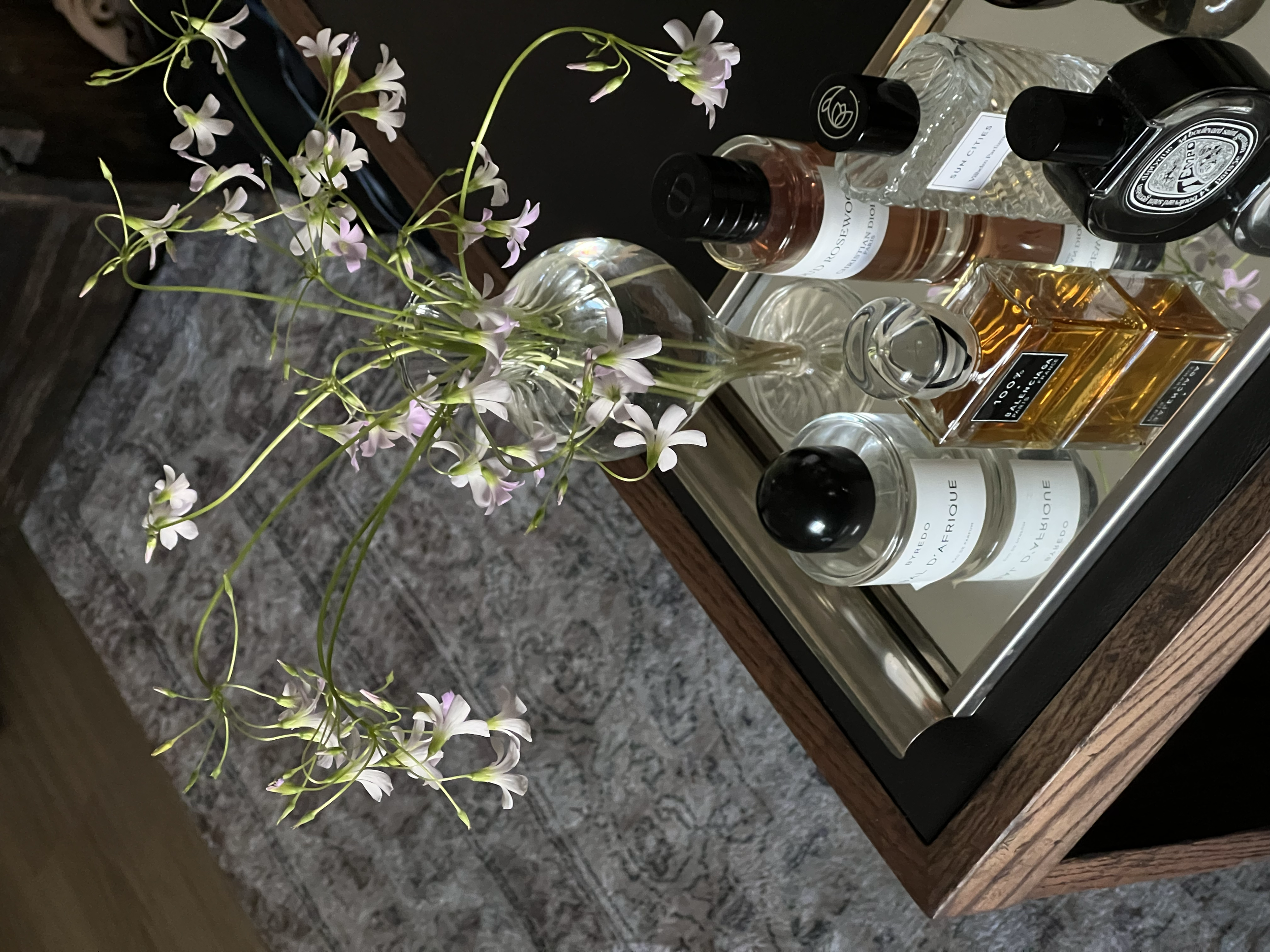 A tray with several bottles of perfume and cologne, a glass vase with pink and white flowers, and a small glass container, placed on a wooden surface.
