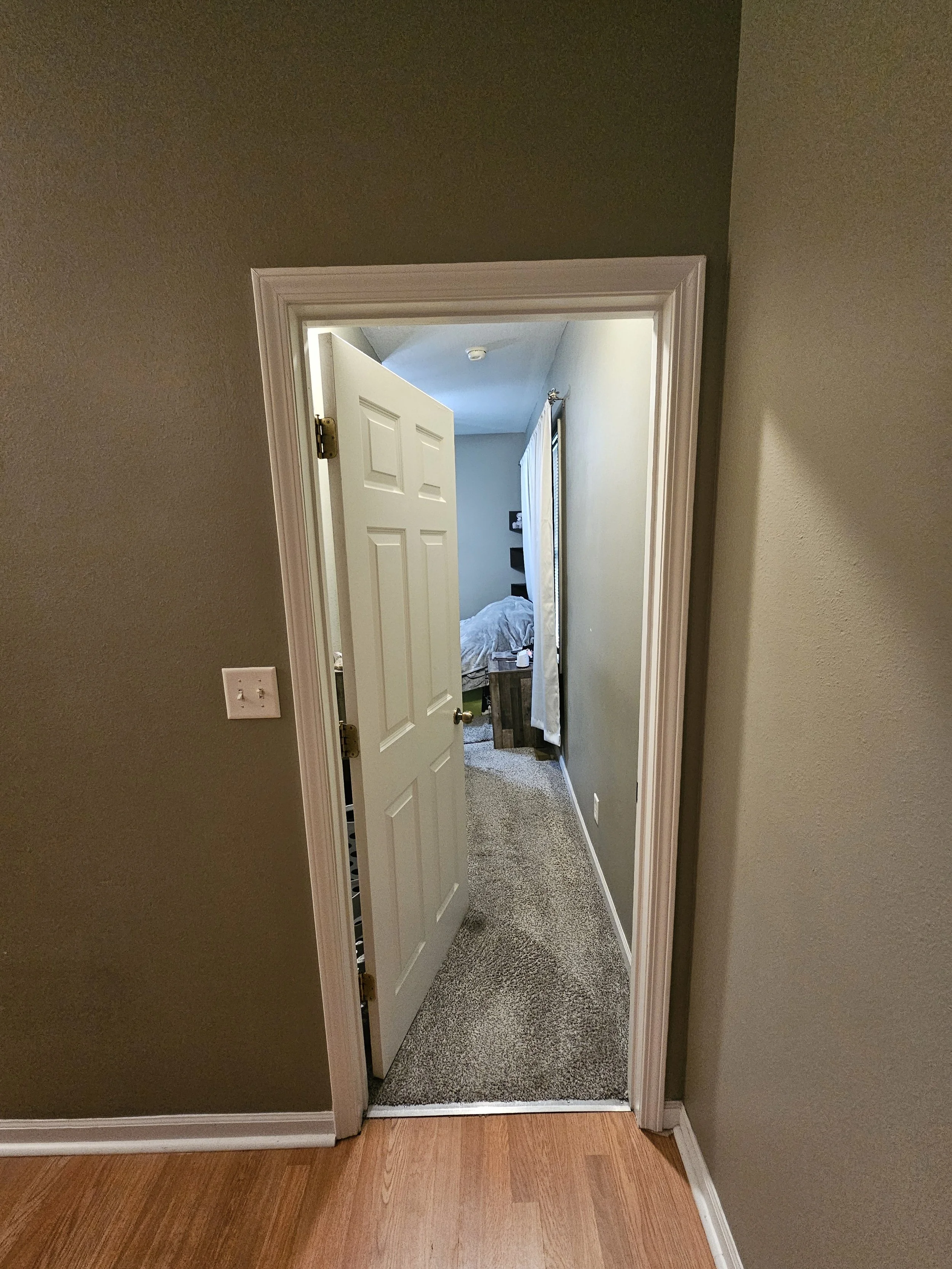 A partially open white door leading into a bedroom with grey walls and a window with curtains, a bed with rumpled sheets, a wooden nightstand with items on it, and a beige carpeted floor.