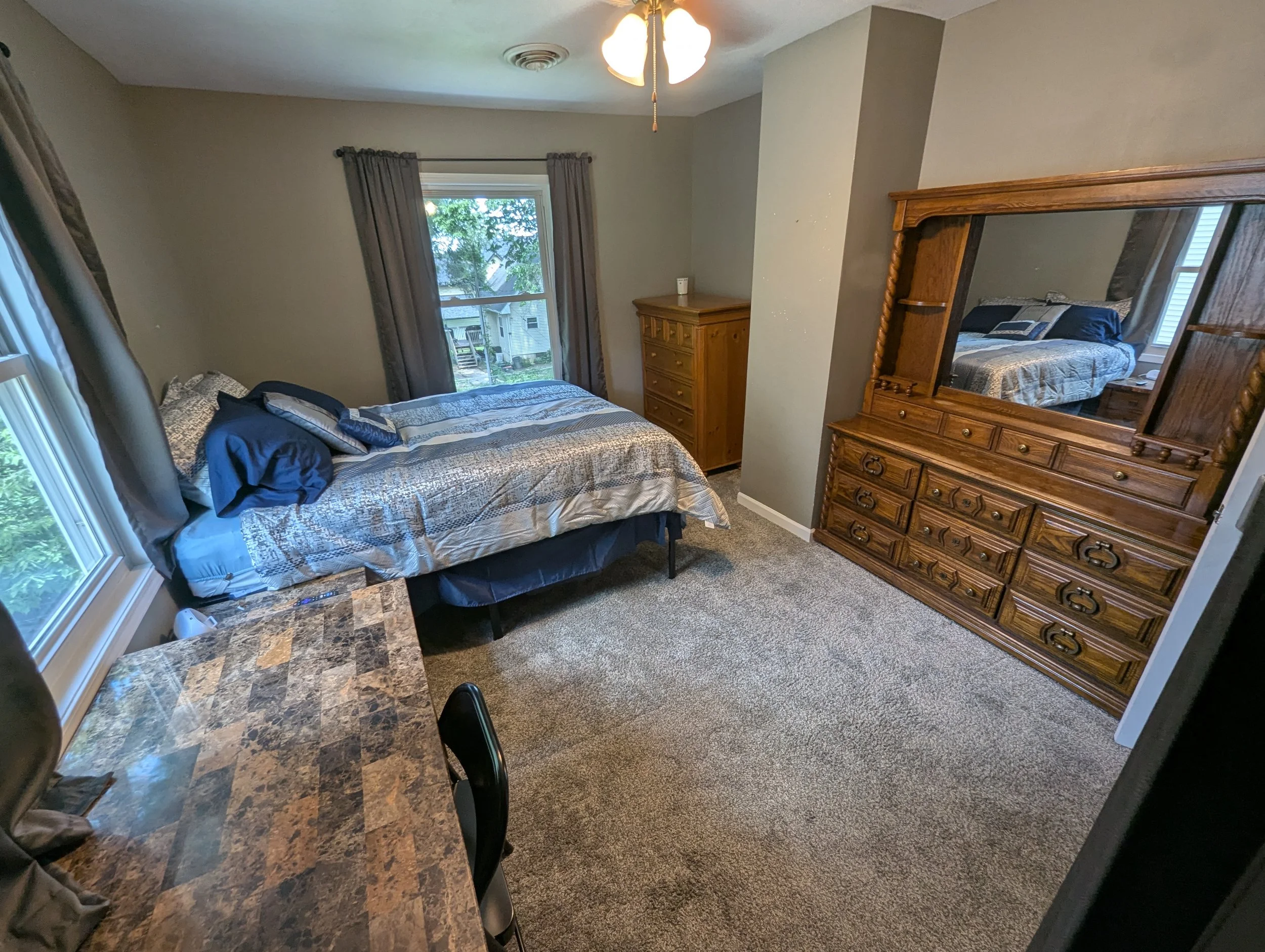 A bedroom with a bed covered in blue and white patterned bedding, a wooden dresser with a mirror, a window with brown curtains, a ceiling fan with light, a small table with a patterned surface, and a carpeted floor.