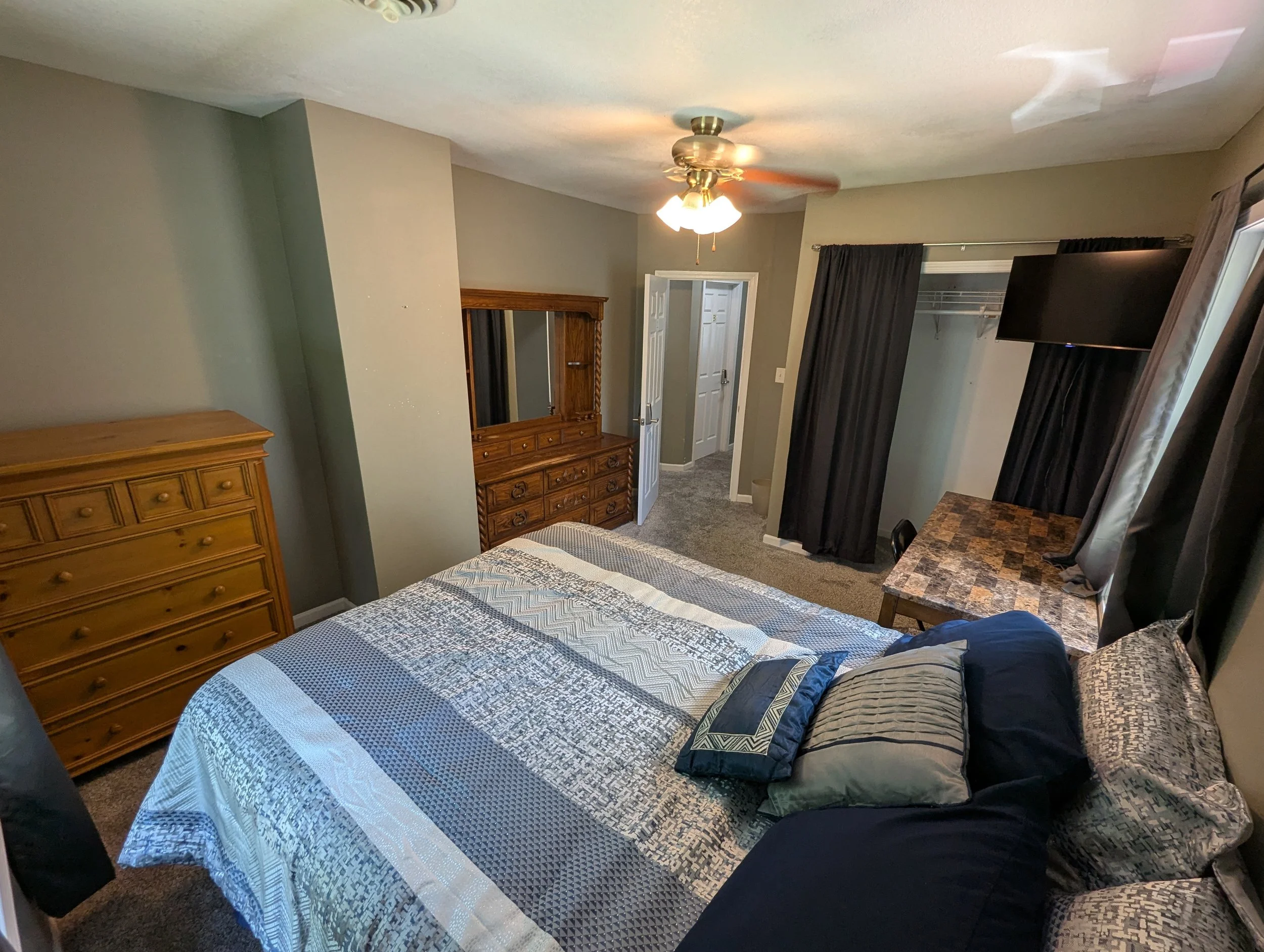 A bedroom with a bed covered in a blue, white, and gray quilt, and multiple pillows. There is a wooden dresser with a mirror, a TV mounted on the wall, and dark curtains over a window. A ceiling fan with lights is turned on.
