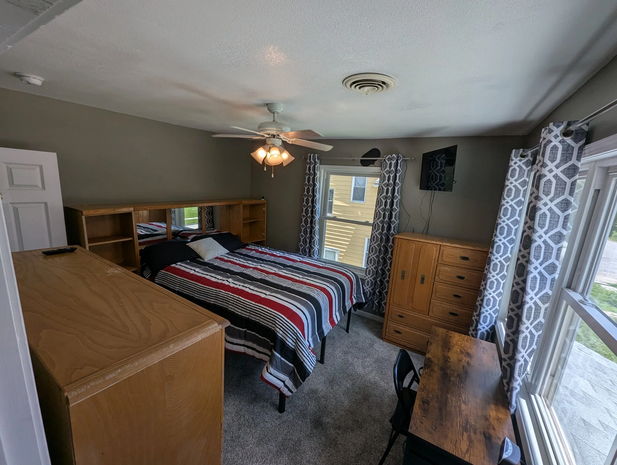 A bedroom with a bed covered in a striped red, black, and white blanket, wooden furniture, patterned curtains, a ceiling fan with lights, and a flat-screen TV mounted on the wall.