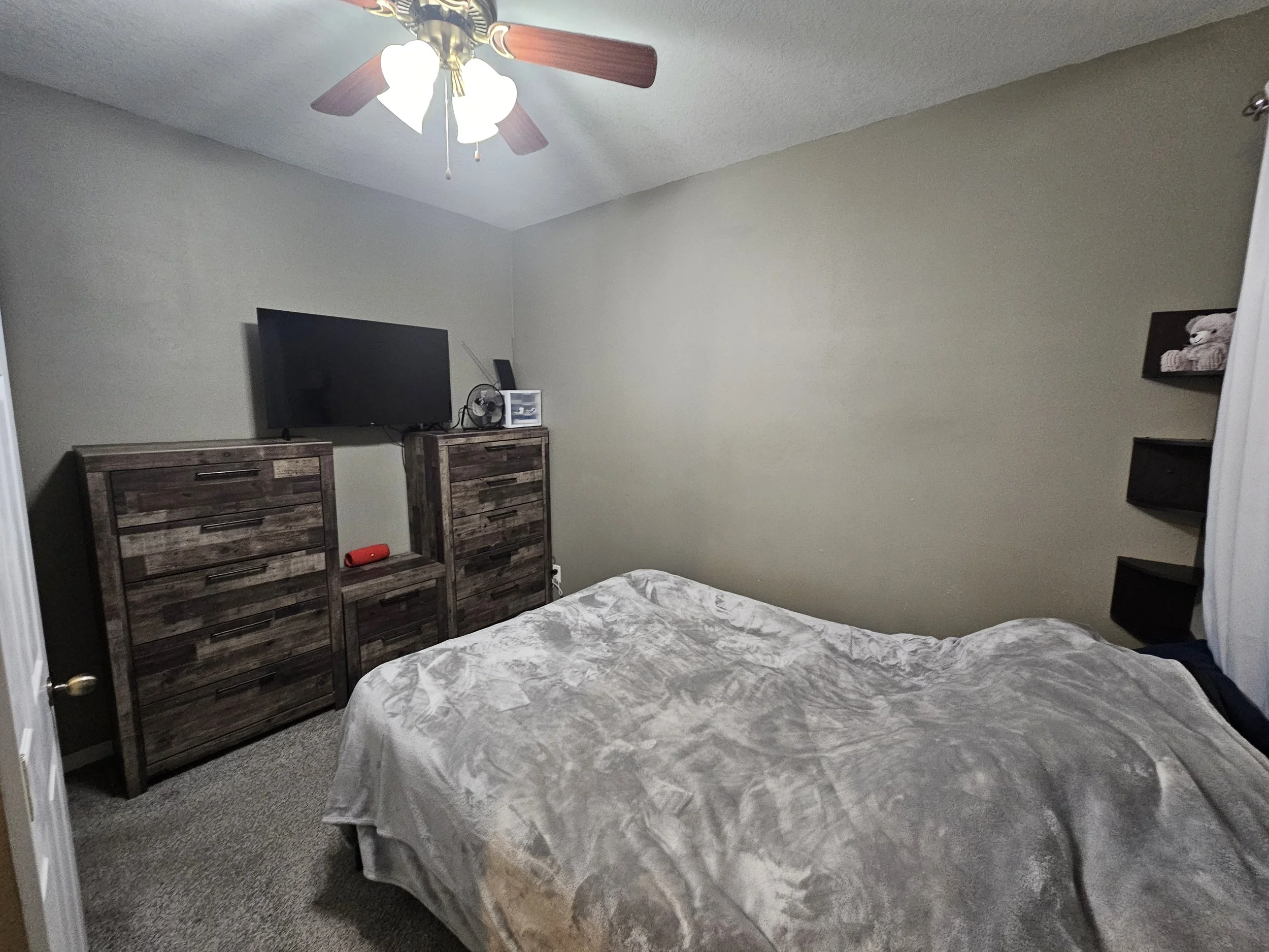 A bedroom with beige walls, a ceiling fan with lights, a flat-screen TV on a rustic wooden dresser, a white storage container, a bed with a patterned comforter, and wall shelves with decorations.