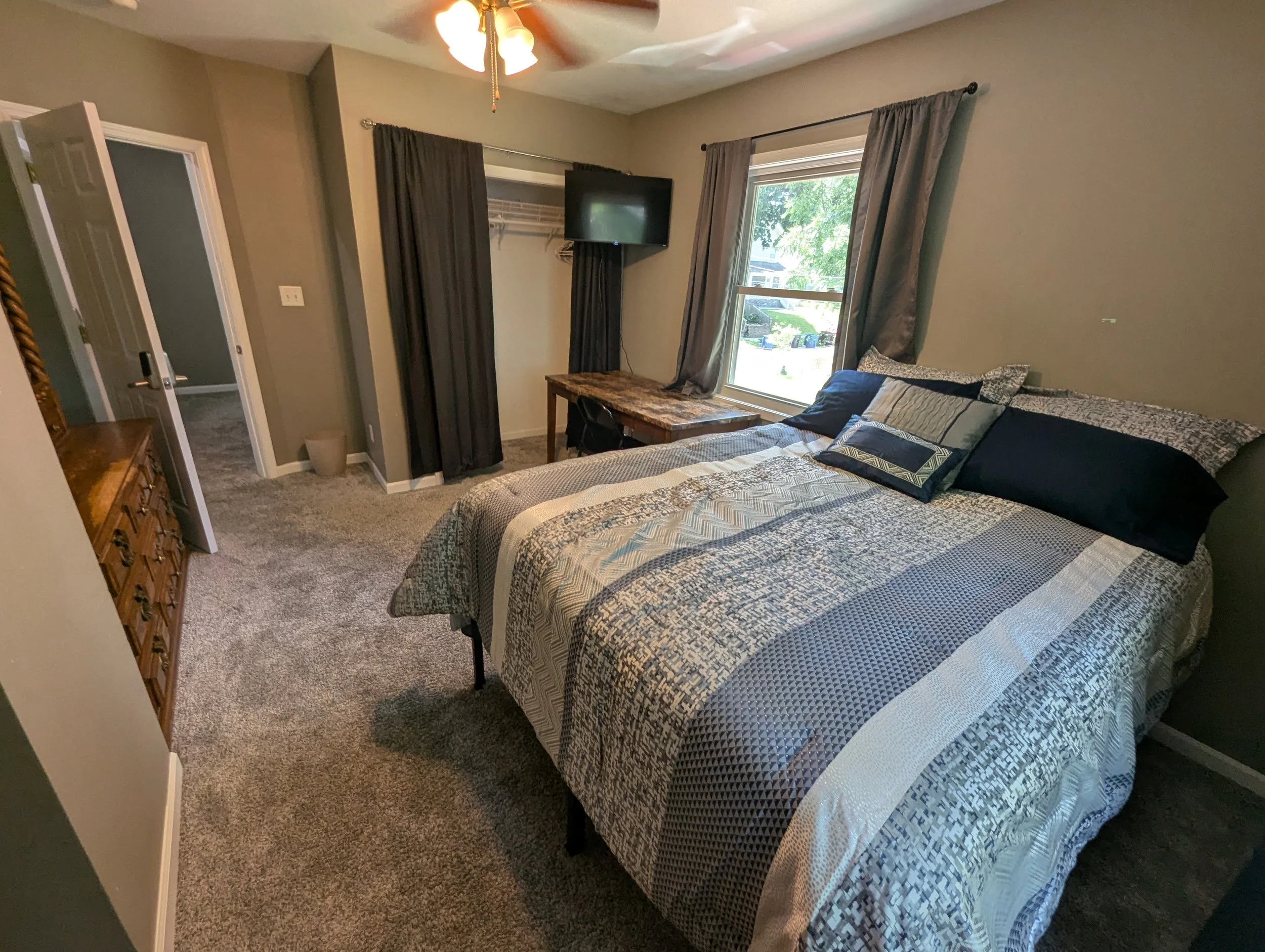 A bedroom with a bed covered in a blue, gray, and white patterned quilt, multiple pillows, a wooden dresser, a window with gray curtains, a flat-screen TV mounted on the wall, a closet with dark curtains, and a ceiling fan with lights.
