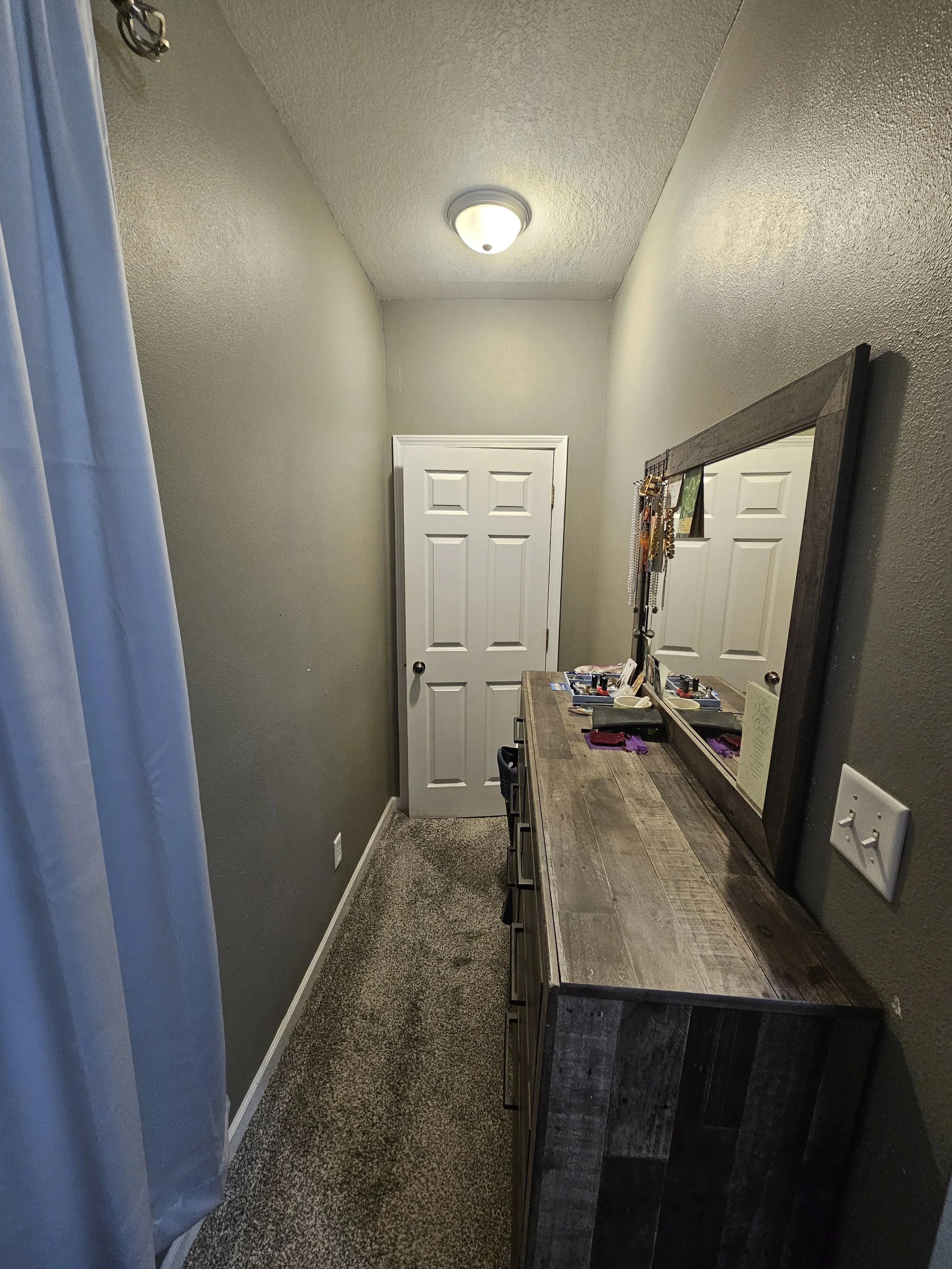 A narrow hallway with beige walls and ceiling, featuring a white door at the end. To the right, there's a wooden dresser with a large mirror, jewelry and personal items on top, and a purple cloth. On the left, part of a white curtain is visible. The 